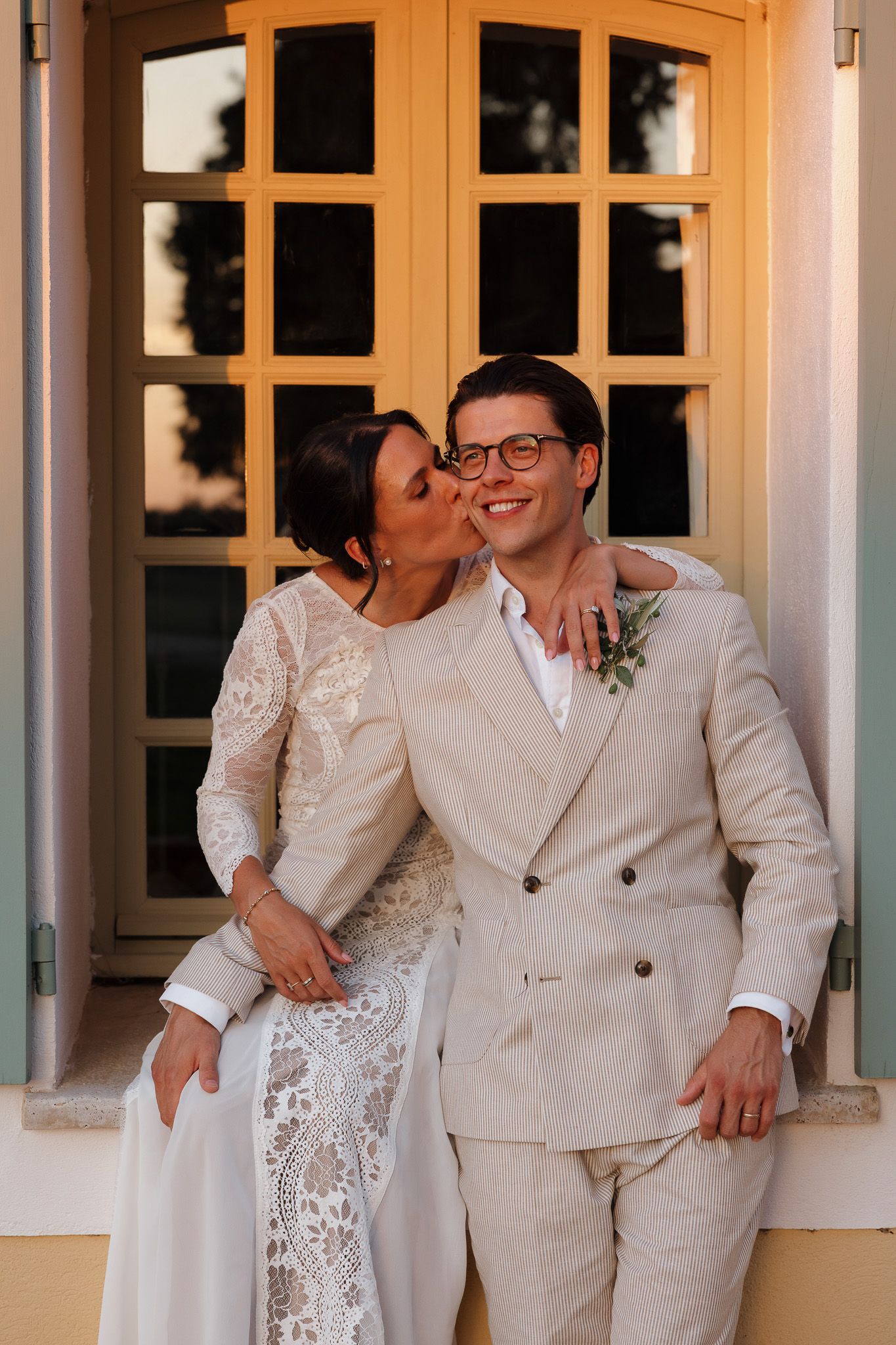Bride in lace gown kisses groom in beige suit on cheek outside golden window at outdoor wedding.