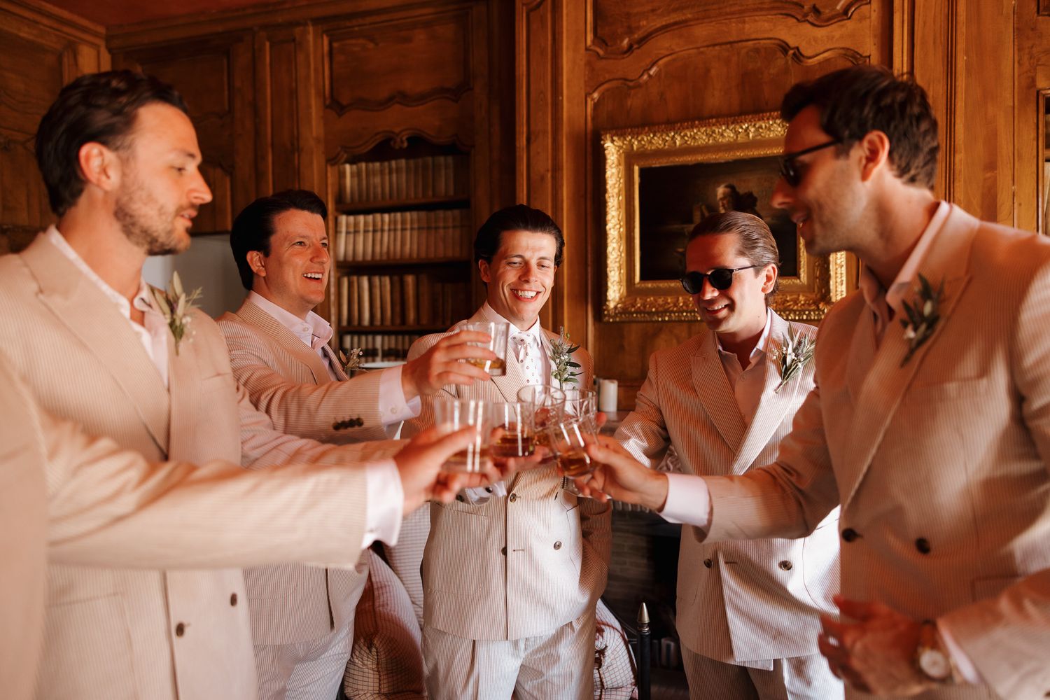Five men in beige suits toasting with champagne glasses in an elegant wood-paneled room during a wedding celebration.