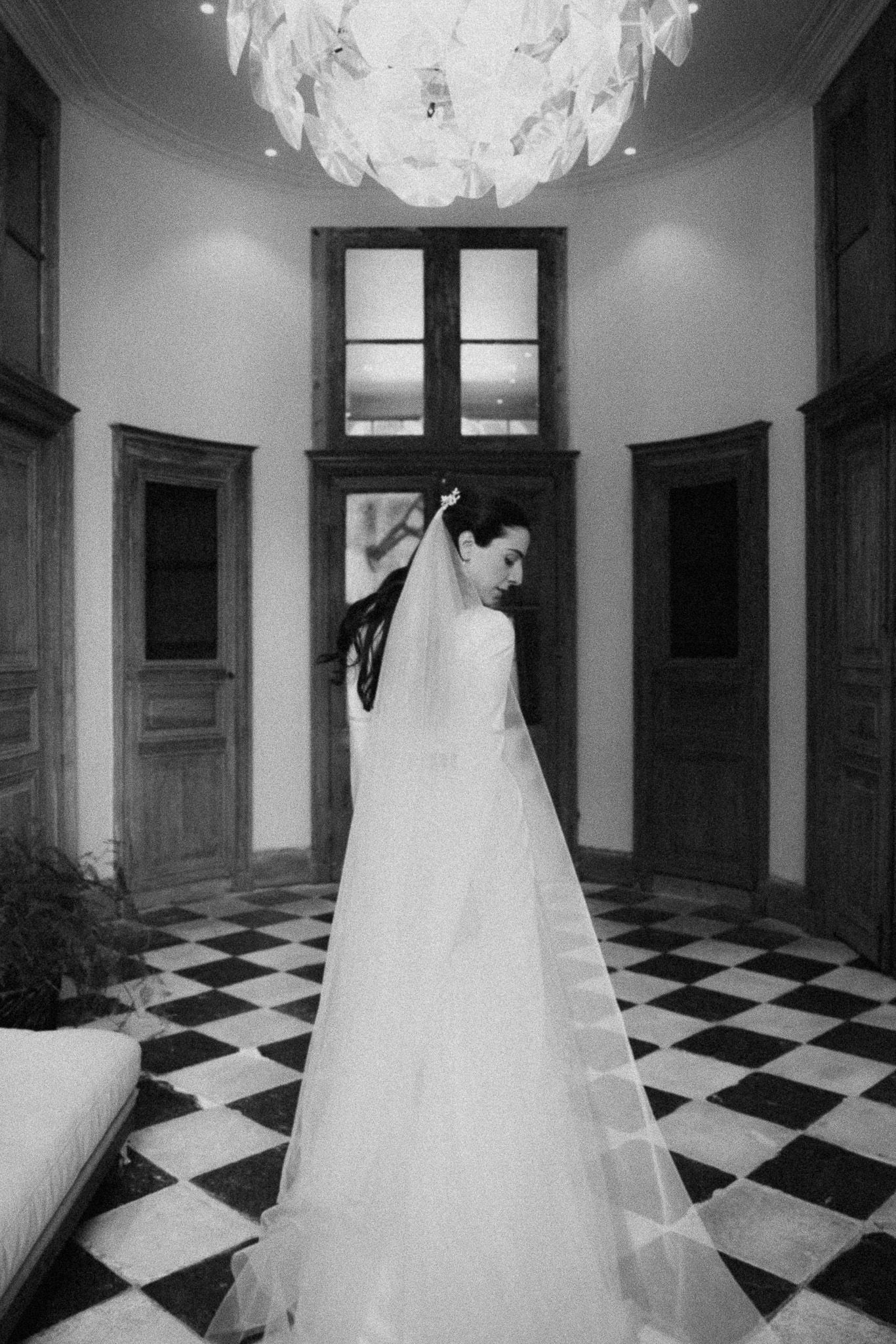 Bride in a white gown and veil stands in a black and white checkered hall beneath a chandelier.