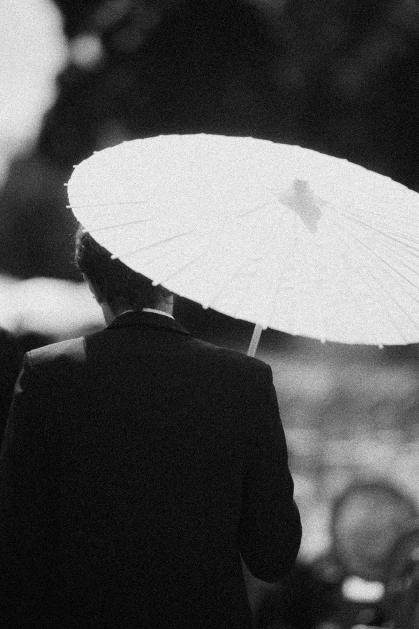 Black and white photo of a person in formal attire holding a white paper parasol umbrella from behind.
