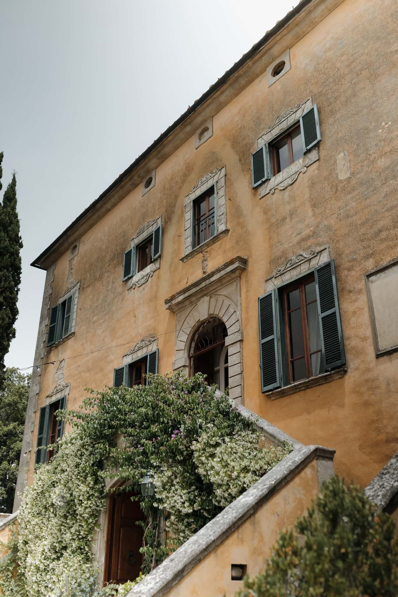 Rustic Italian villa exterior with climbing white flowers and weathered beige stucco walls against a bright sky.
