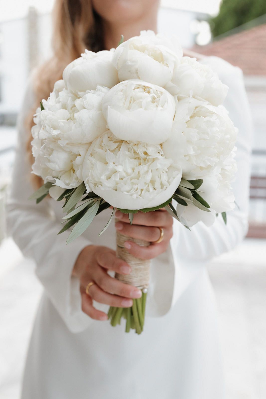 A bride holds a classic white peony bouquet against her wedding dress in an elegant outdoor setting.