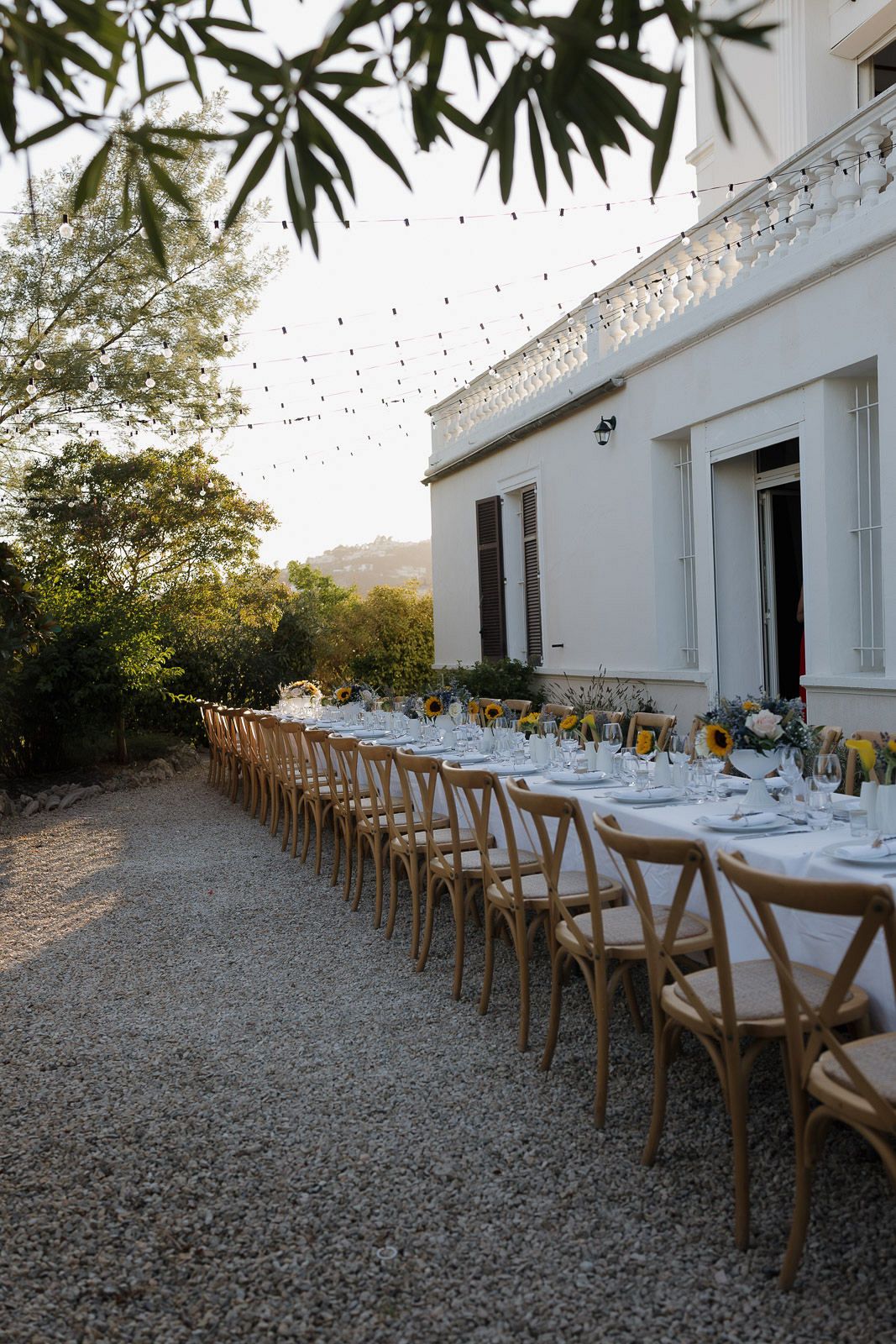 A long outdoor dining table with wooden chairs set for an elegant dinner party under string lights at sunset.