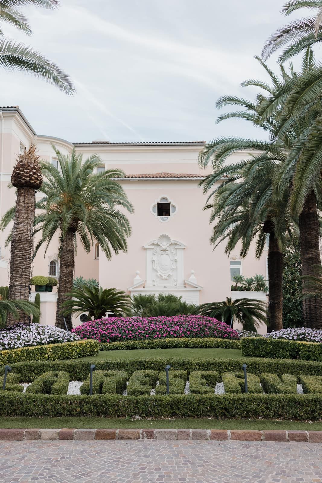 Elegant landscaping with manicured hedges and palm trees line a brick pathway leading to a Mediterranean-style building.