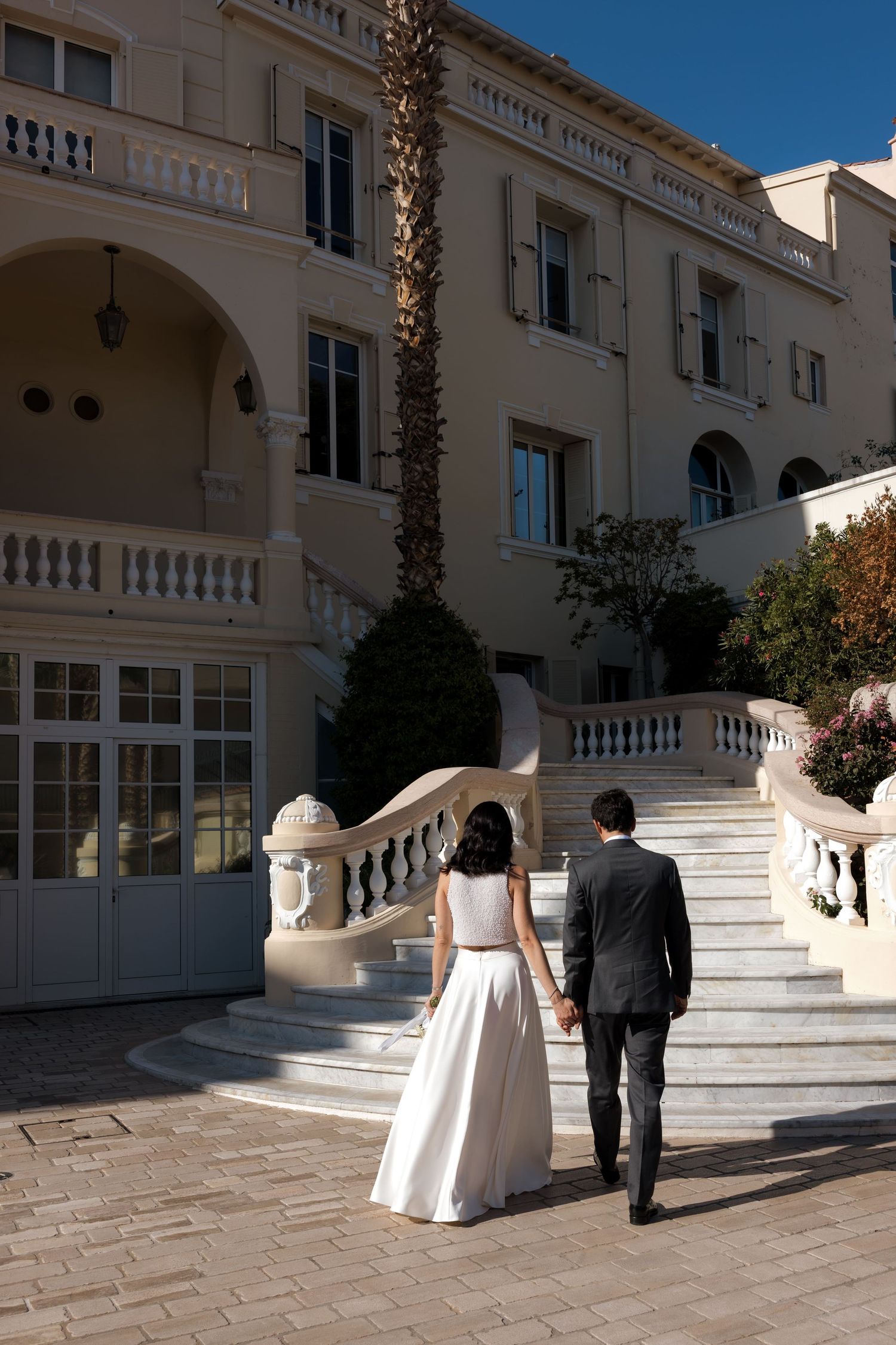 Bride and groom holding hands walking toward elegant white mansion with grand staircase on sunny wedding day.