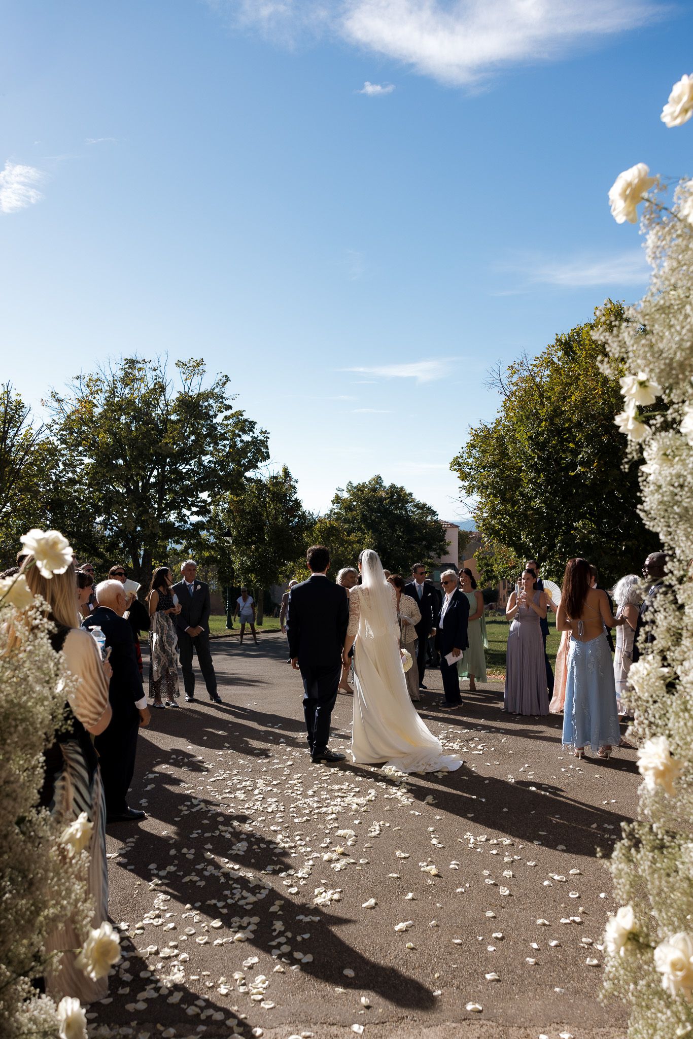 Bride and groom walk down petal-lined outdoor aisle flanked by white floral arrangements and wedding guests under blue sky.