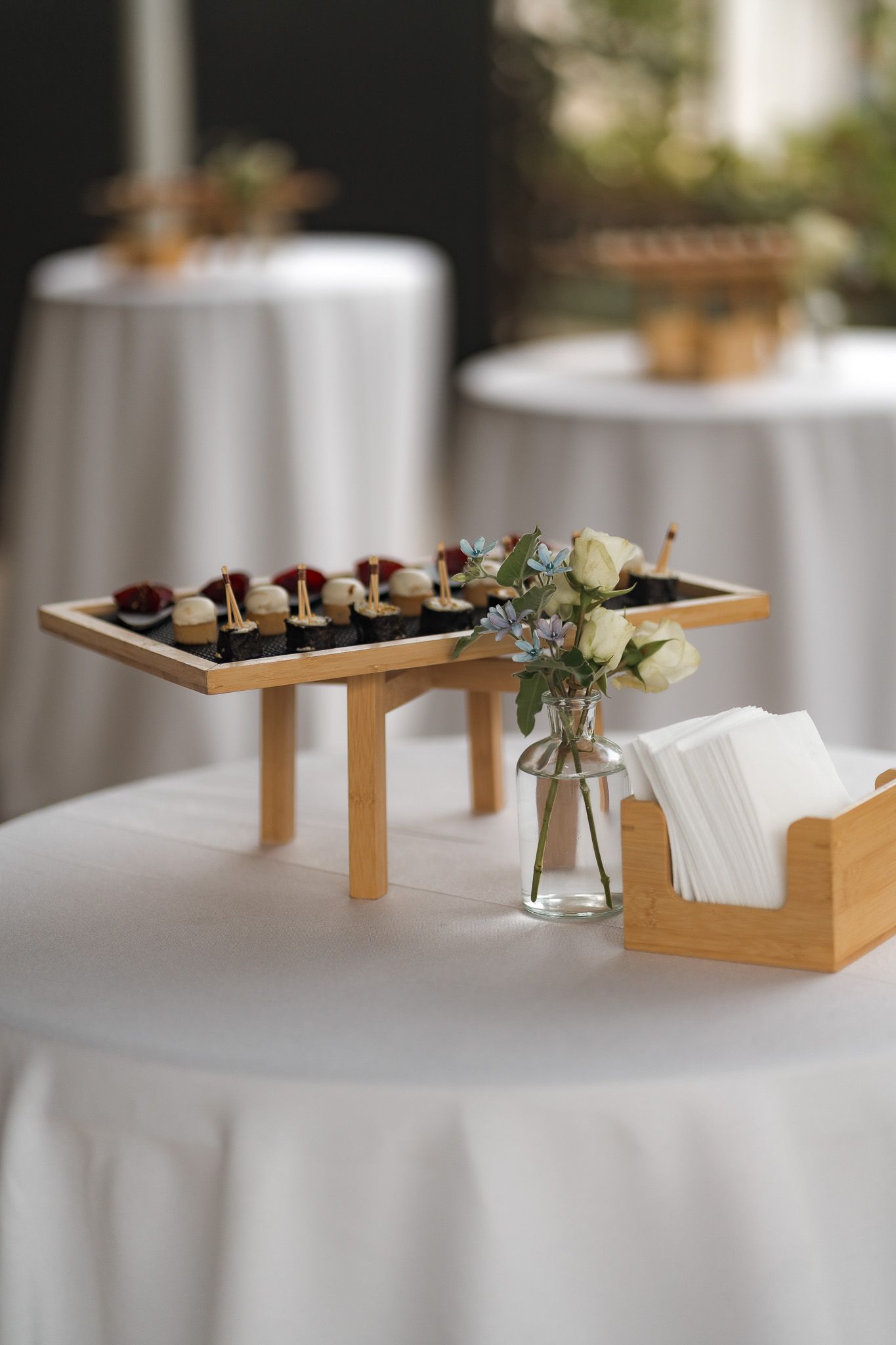 Elegant catering display with appetizers on a wooden riser, floral vase, and napkin holder on a white tablecloth.