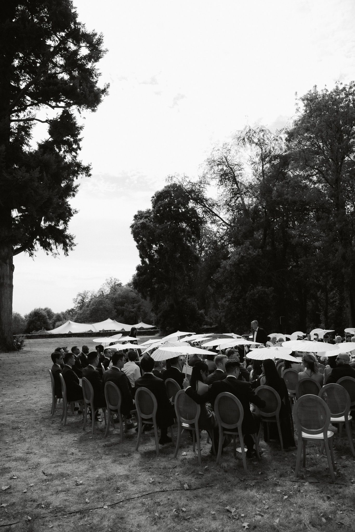 Black and white outdoor wedding ceremony with guests seated in chairs under parasols surrounded by tall trees.