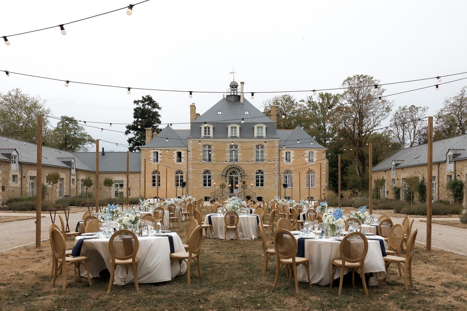 Elegant outdoor wedding reception set up in front of a French chateau with string lights and wooden chairs.