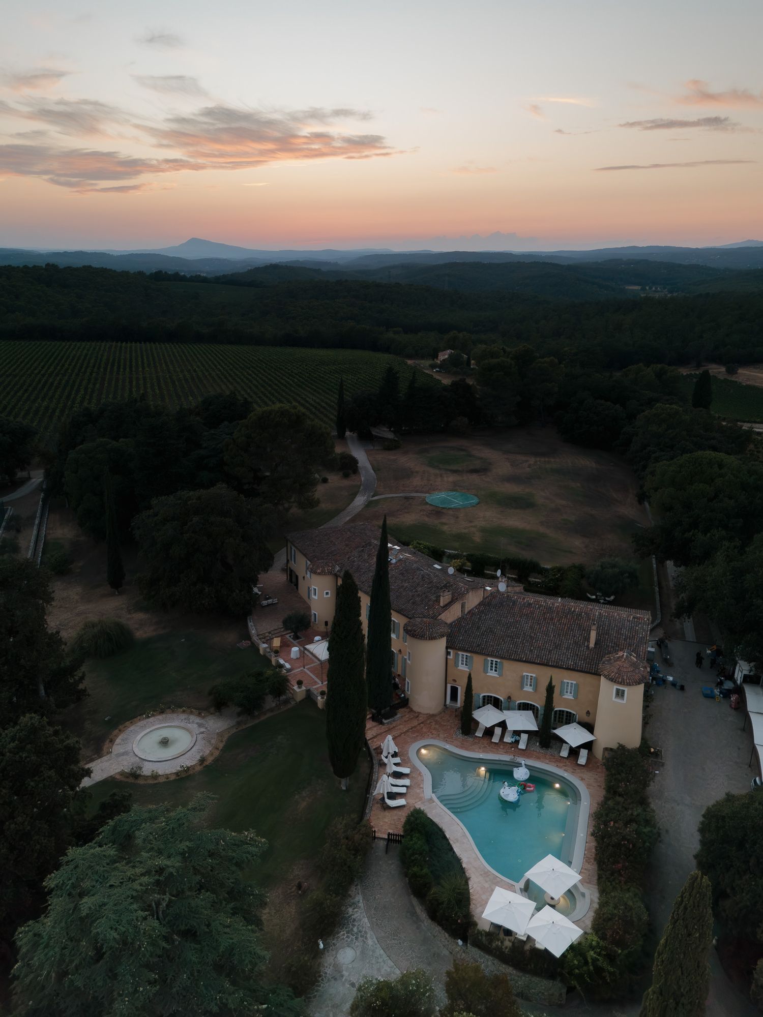 Aerial drone view of a luxury French chateau with pool at sunset, surrounded by vineyards and rolling hills.