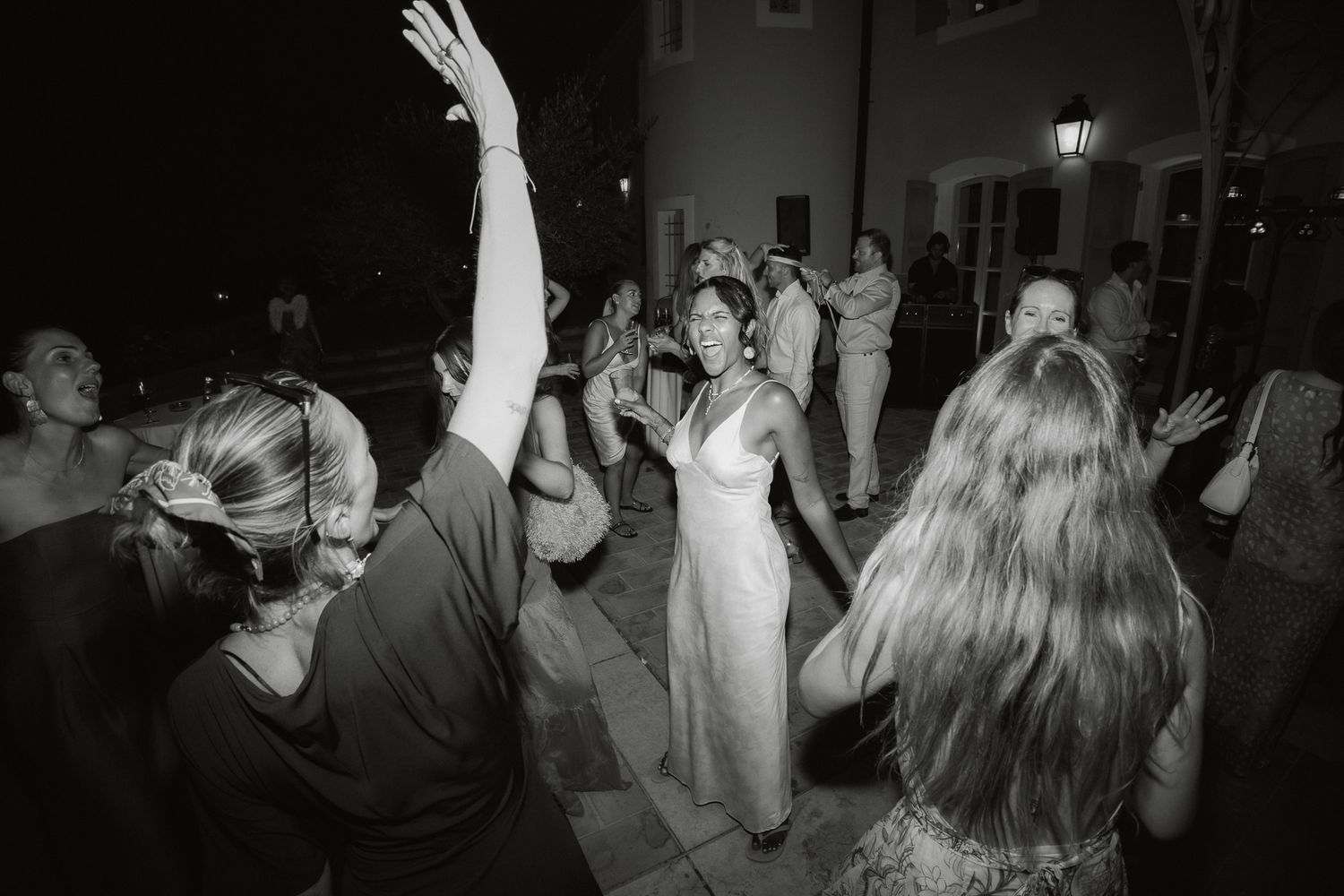 Black and white photo of wedding guests dancing outdoors at night, bride in white dress at center.