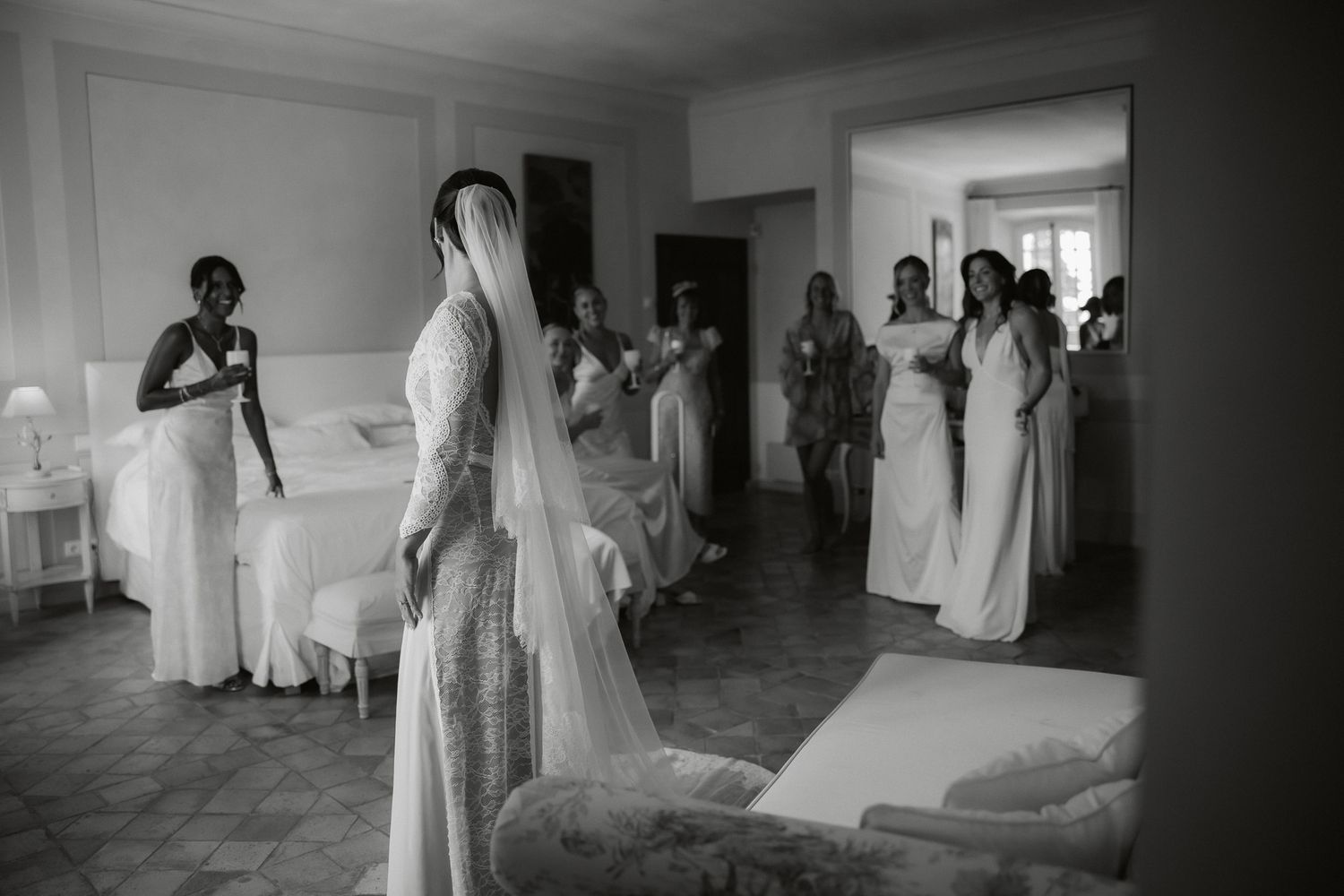 Black and white photo of a bride in a lace gown and veil surrounded by bridesmaids in white dresses before the wedding.