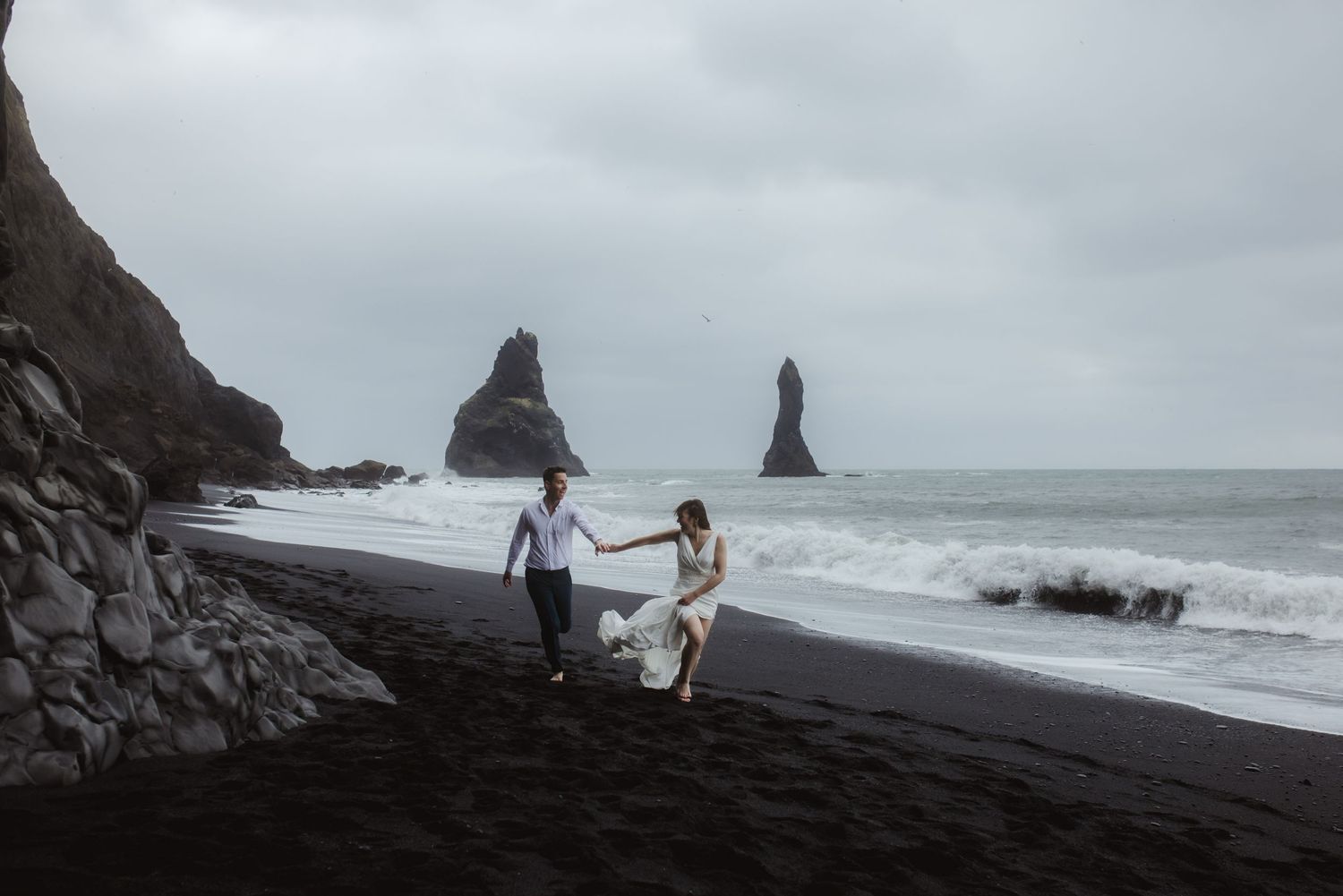Couple enjoys romantic walk along black sand beach in Iceland with dramatic rocky coastline and ocean waves.