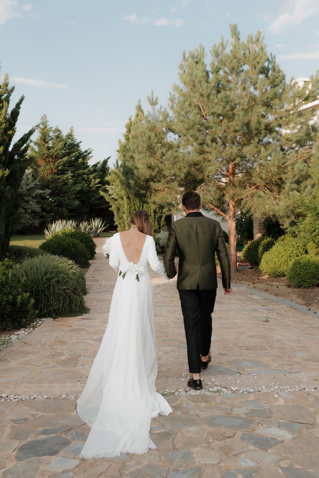 A couple in wedding attire walks along a stone pathway surrounded by pine trees and greenery.