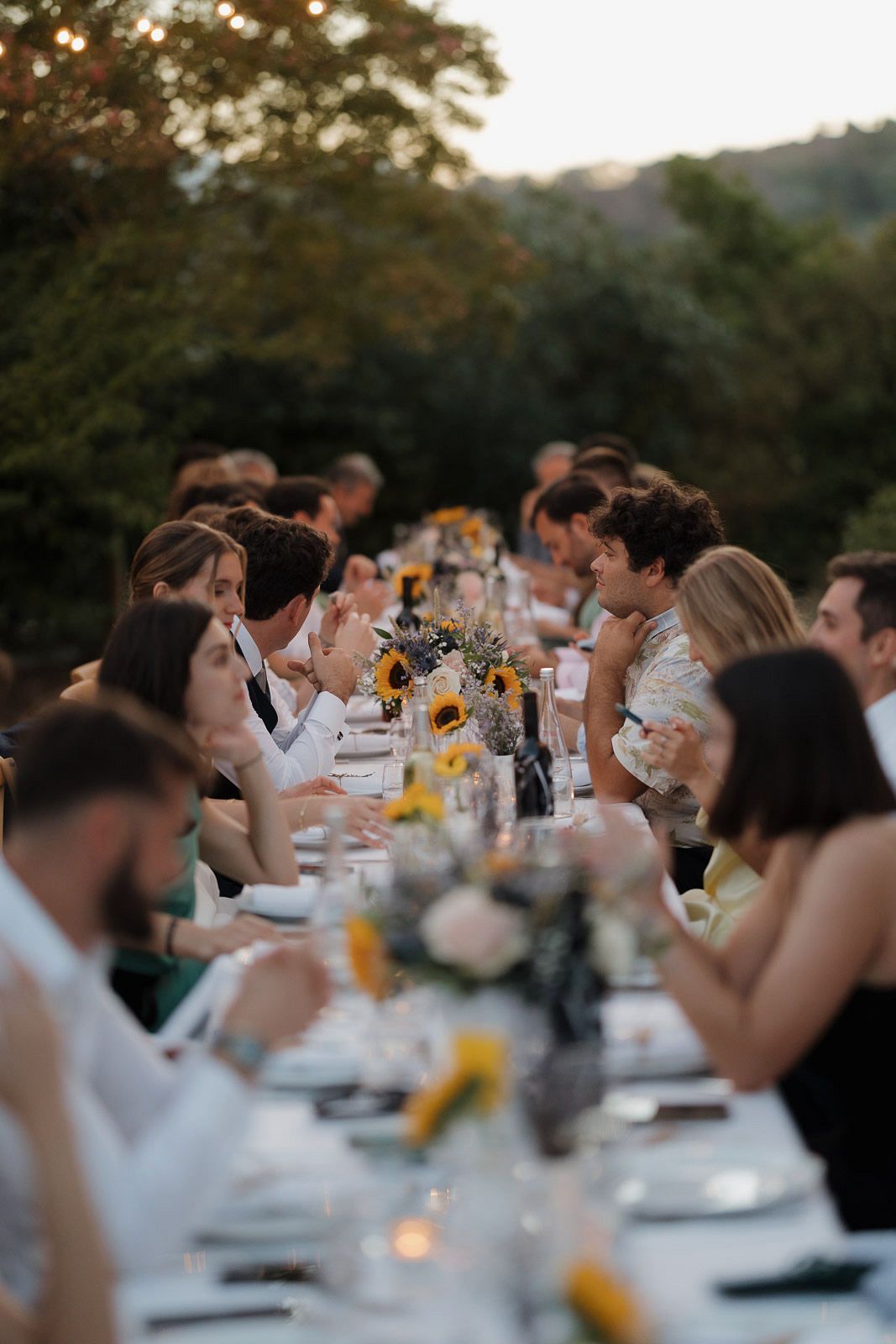 Wedding guests enjoying an outdoor reception dinner at long tables decorated with sunflowers at sunset.