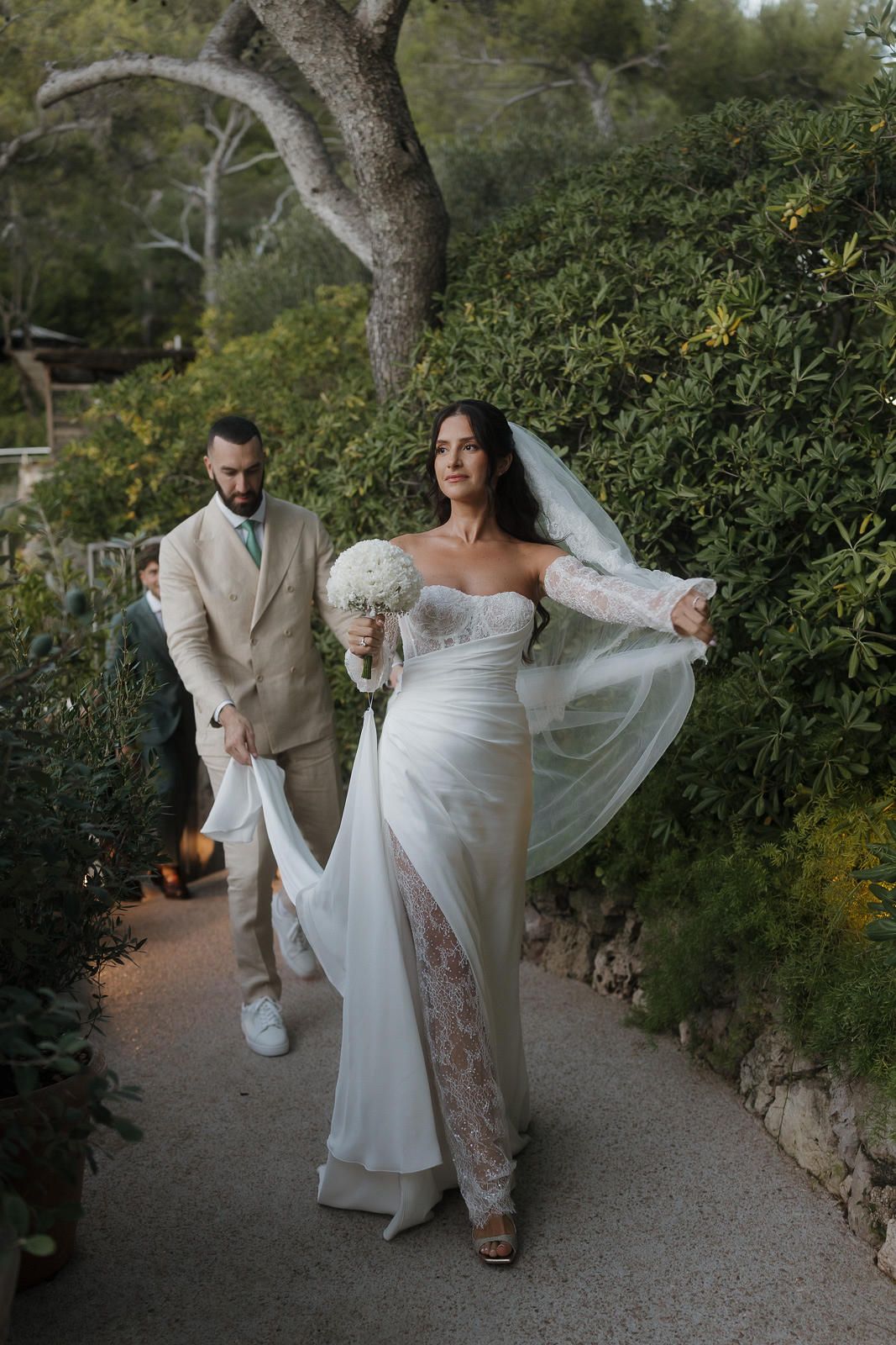 A couple in wedding attire walks playfully along a garden path surrounded by lush greenery.