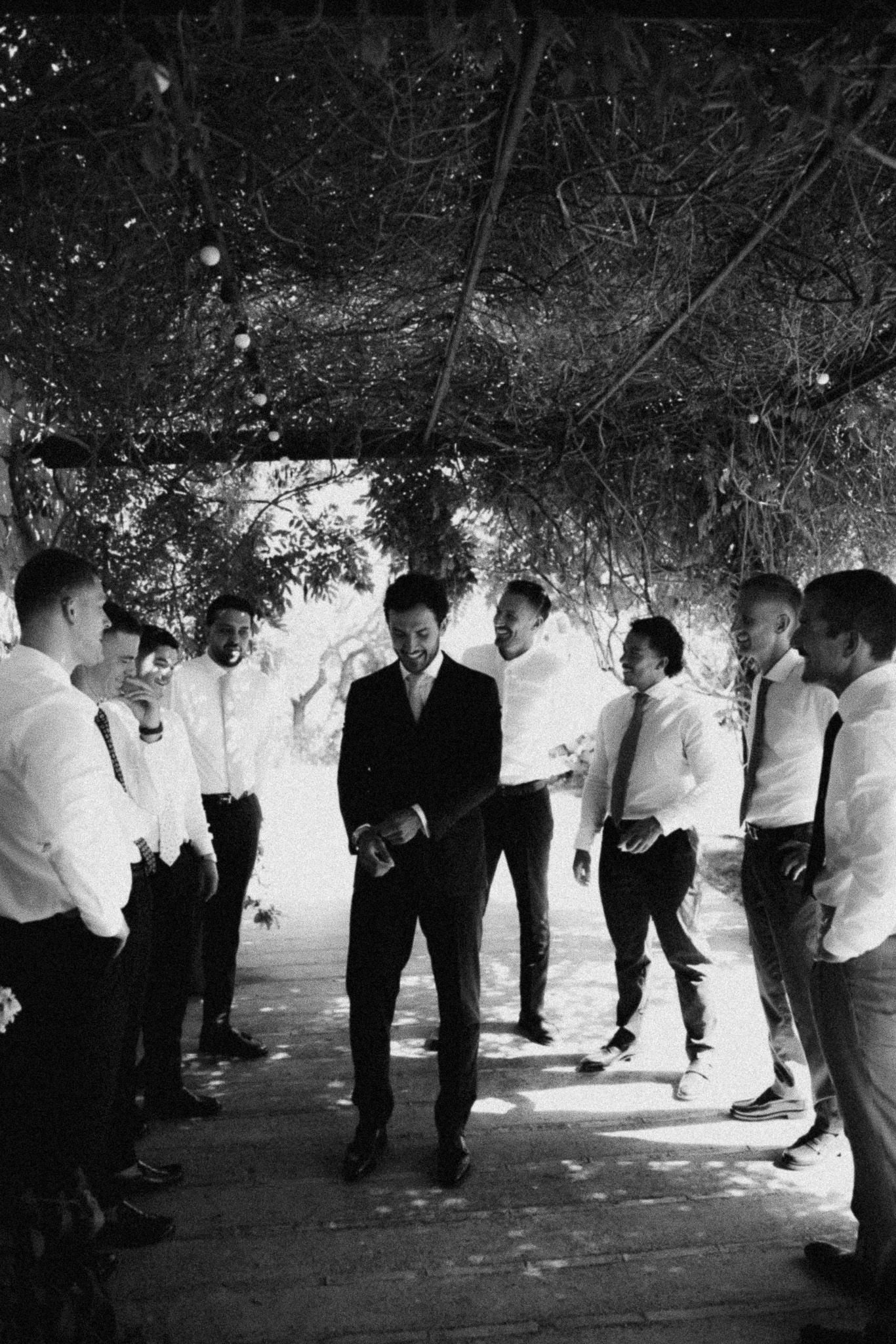Black and white photo of a groom in a suit surrounded by groomsmen in white shirts outdoors.