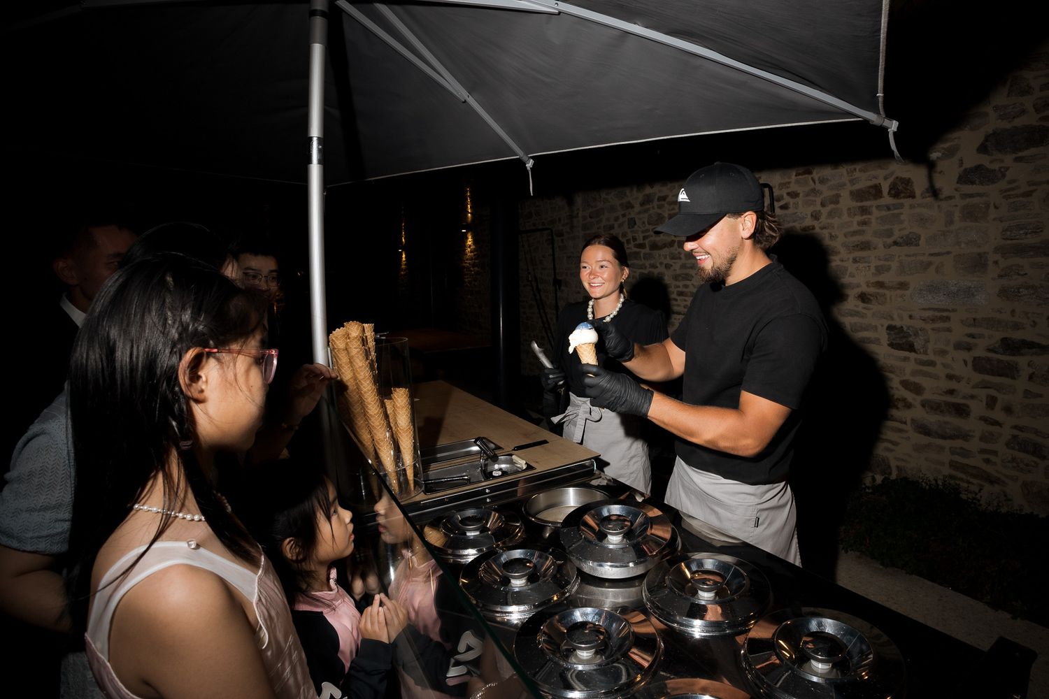 Night market vendor serves ice cream cones to customers at an outdoor food stall with metal bowls on display.