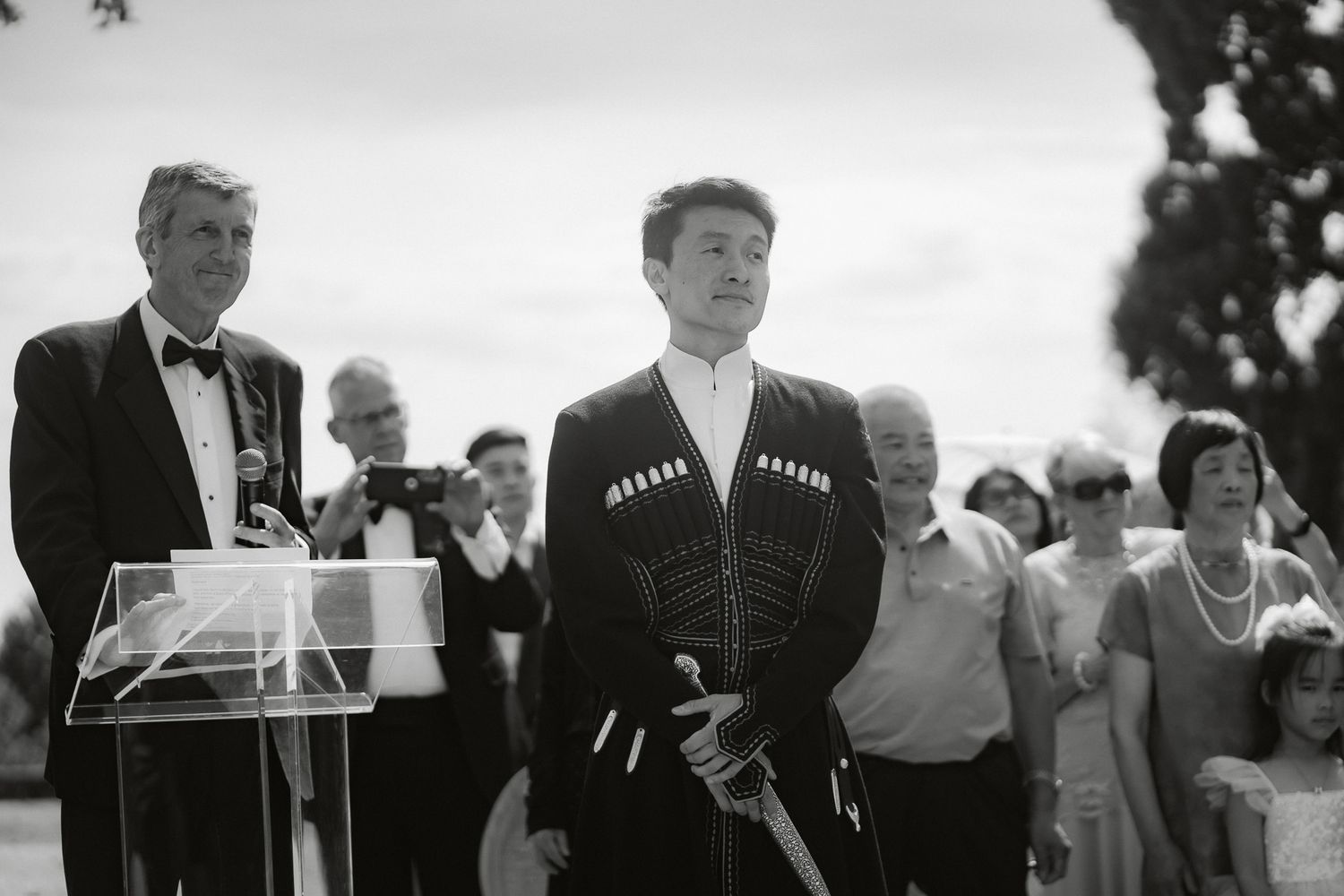 Young man in traditional ceremonial attire stands proudly at an outdoor formal event beside a podium.