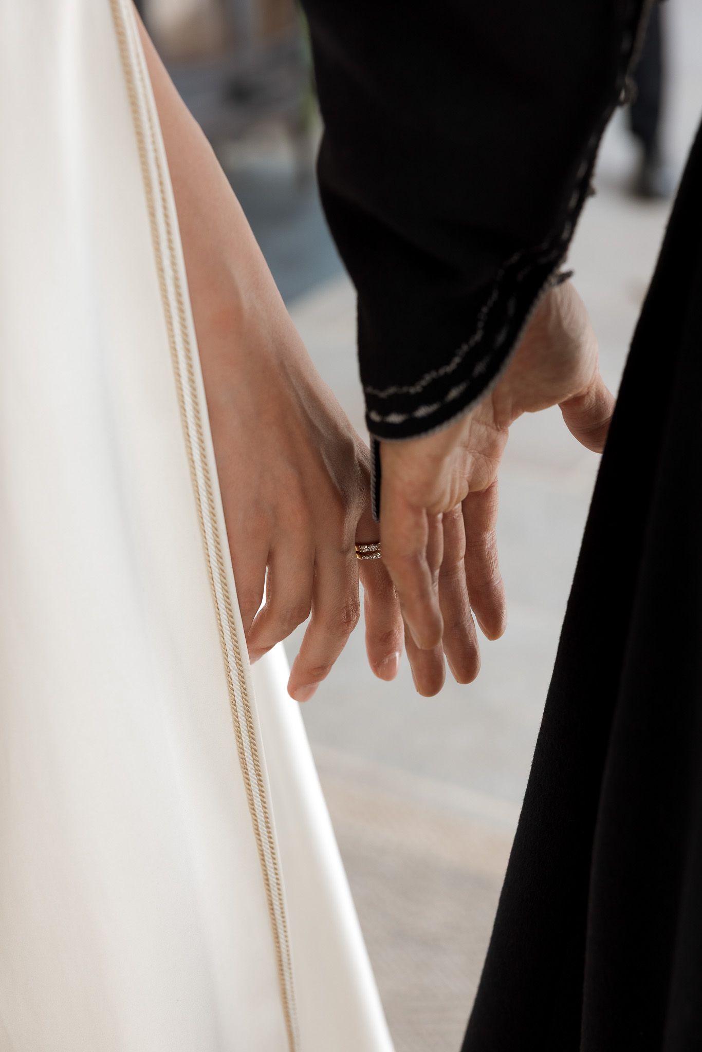 Two hands gently touching, one wearing a ring, against a white wedding dress and black garment.