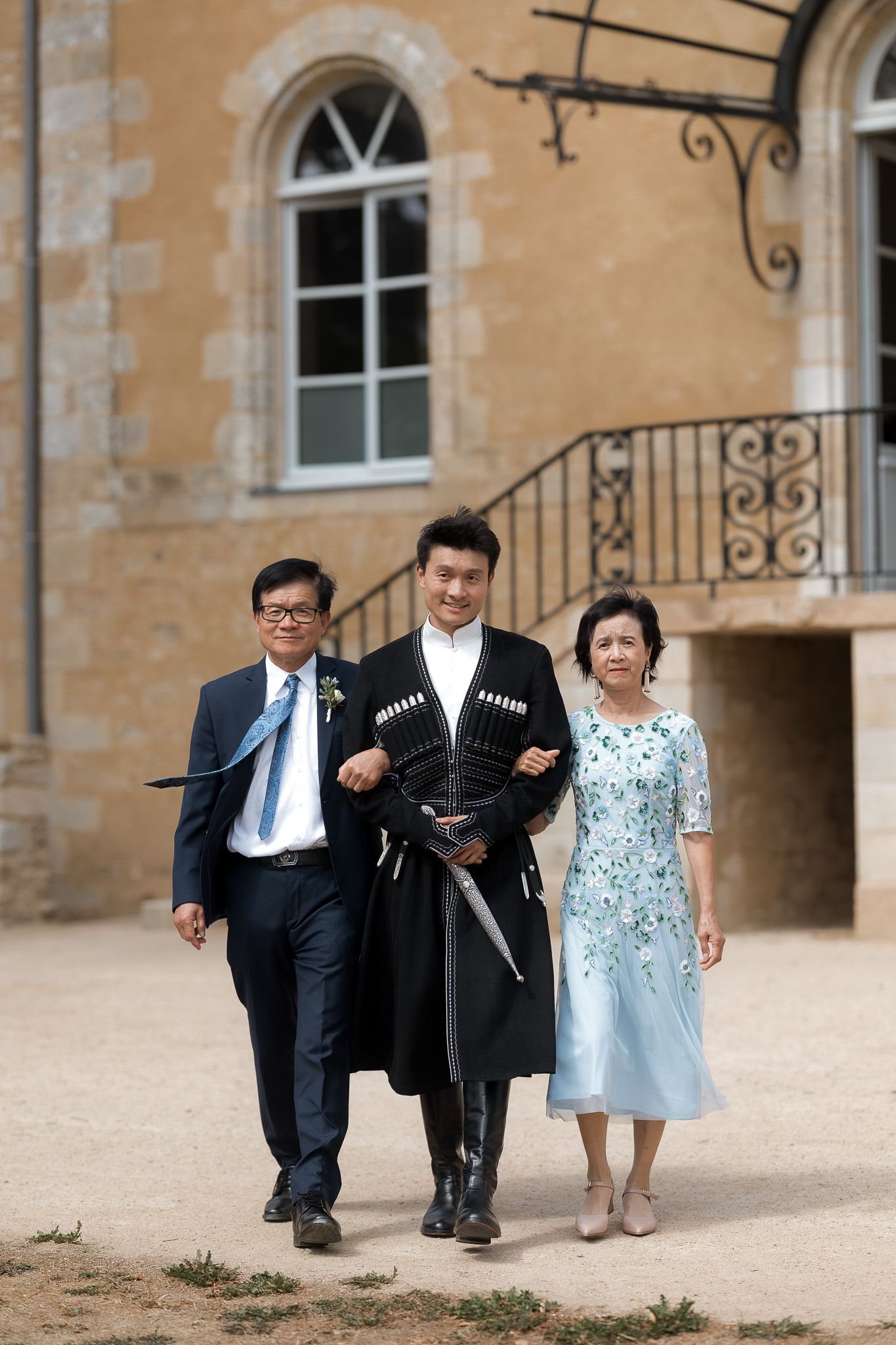 Three people in formal attire pose together outside a stone building, with one man in traditional black ceremonial dress.