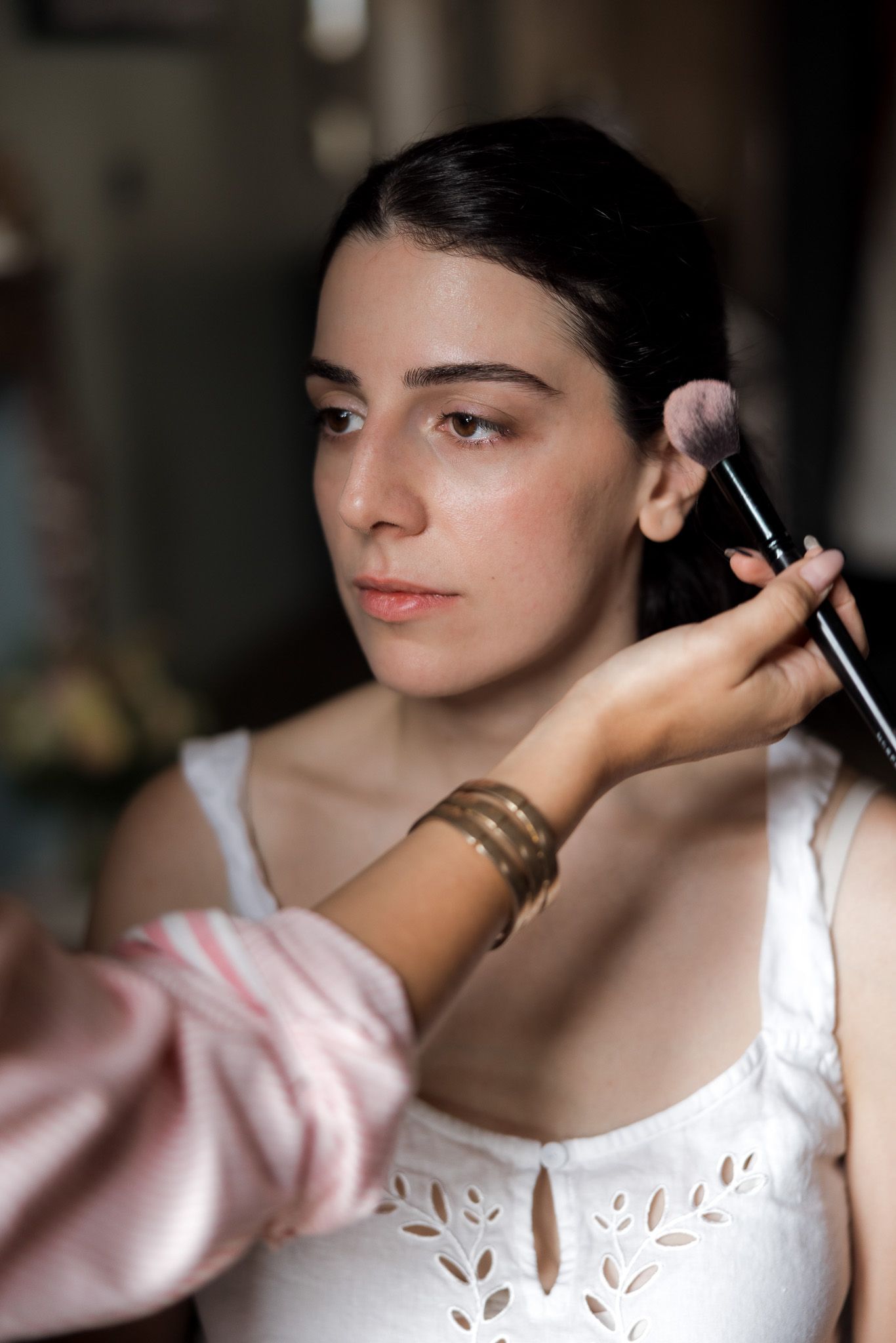 A makeup artist applies blush to a young woman in a white dress using a brush, preparing her for a special occasion.