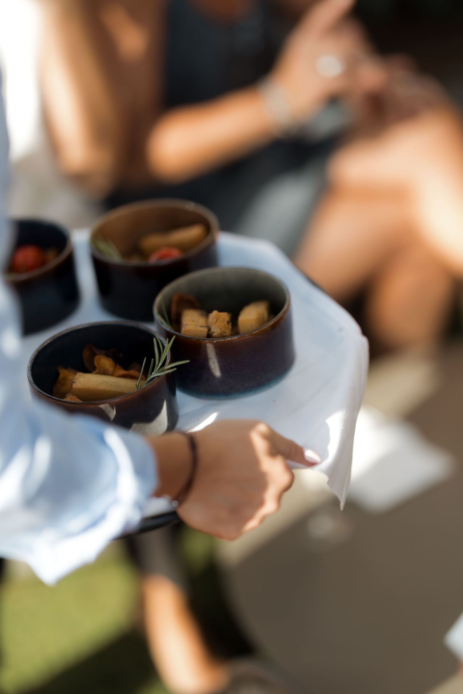 Waiter serving small dark bowls of appetizers on a white cloth tray at an outdoor catering event.