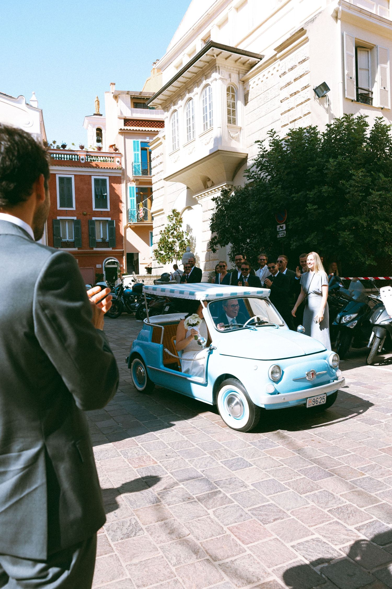 A light blue vintage Fiat 500 drives through a sunny European plaza surrounded by elegant buildings and well-dressed guests.