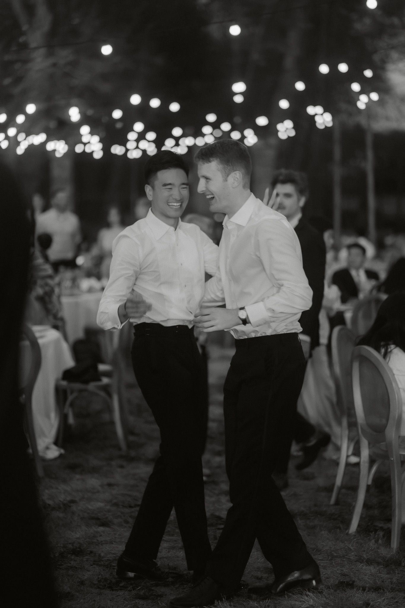 Two grooms share a joyful dance under string lights at their outdoor wedding reception in black and white photo.