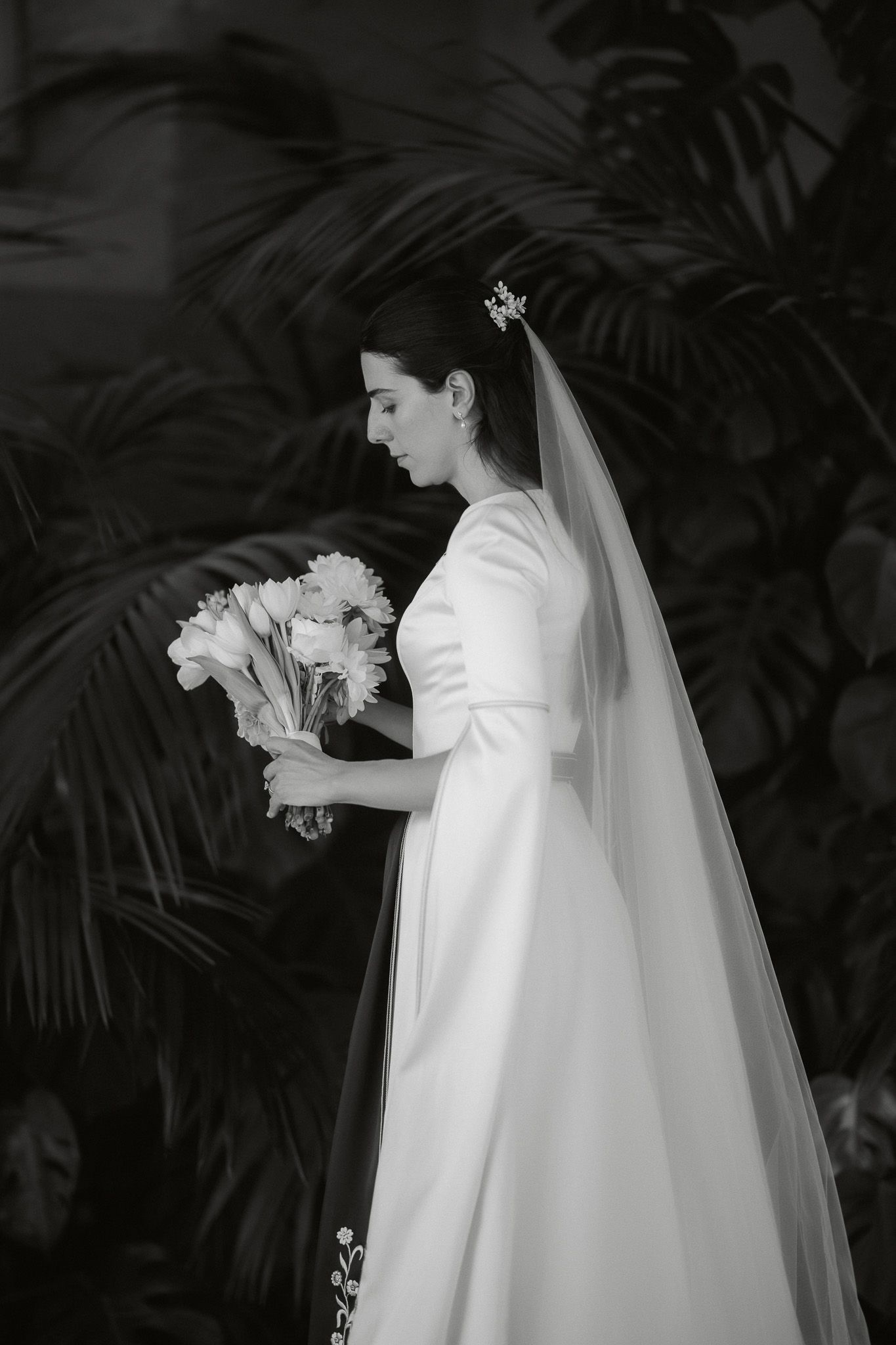 Elegant bride in white gown and veil holds floral bouquet, black and white portrait with tropical foliage backdrop.