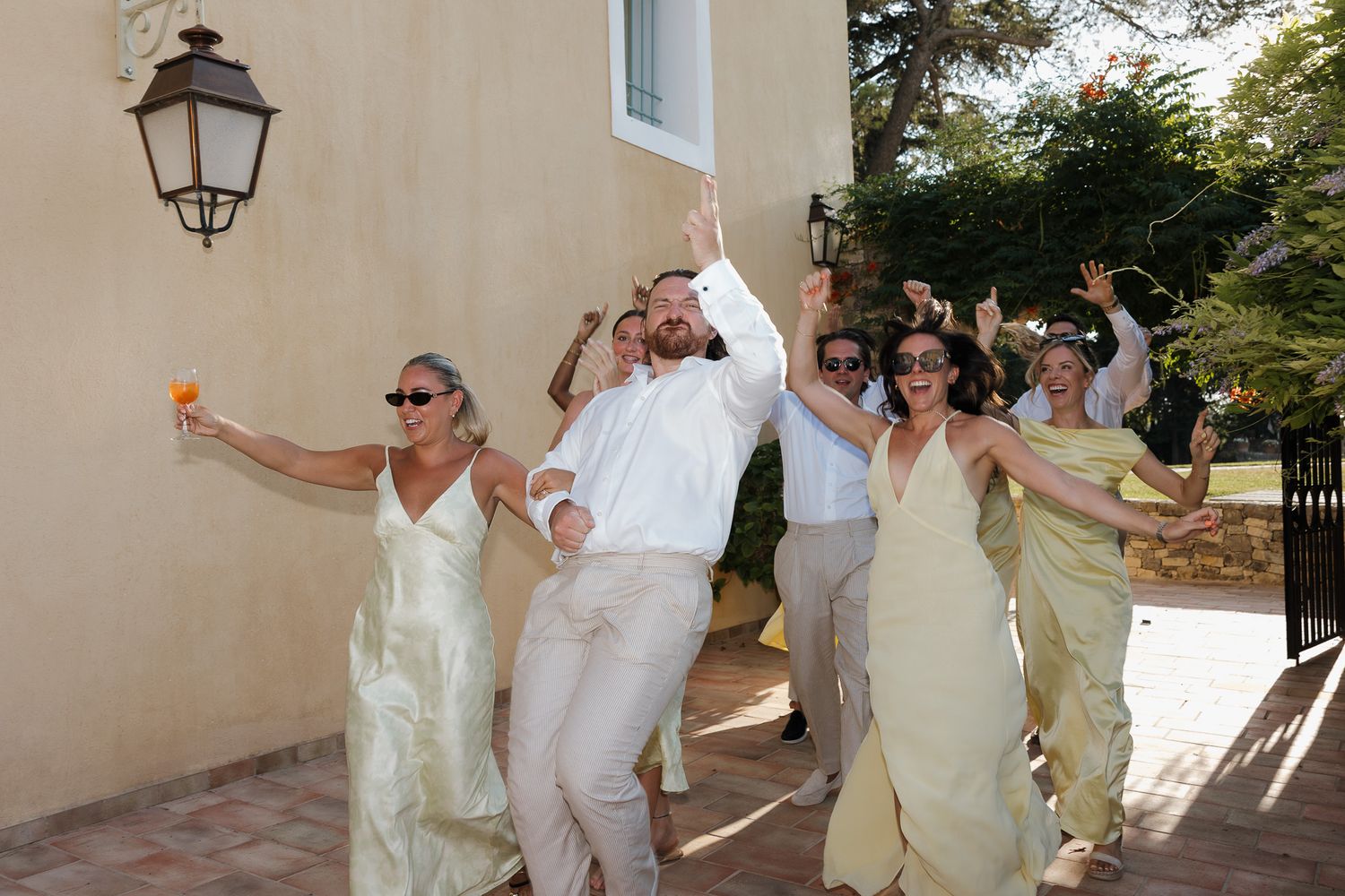 Joyful wedding party celebrating outdoors in sunlight, bridesmaids in gold dresses and groomsmen cheering near a stone building.
