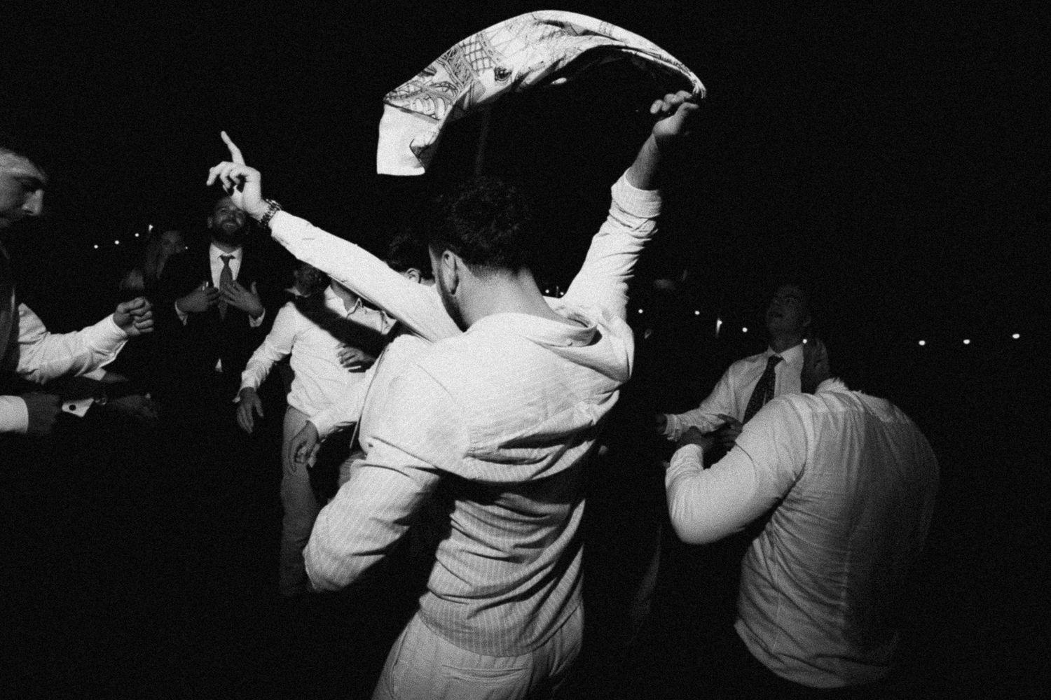 Black and white photo of people dancing energetically at a nighttime outdoor event or wedding celebration.
