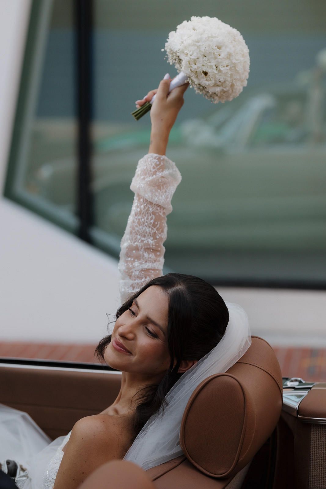A bride in a white lace dress reclines on a brown leather seat while holding up a white floral bouquet.