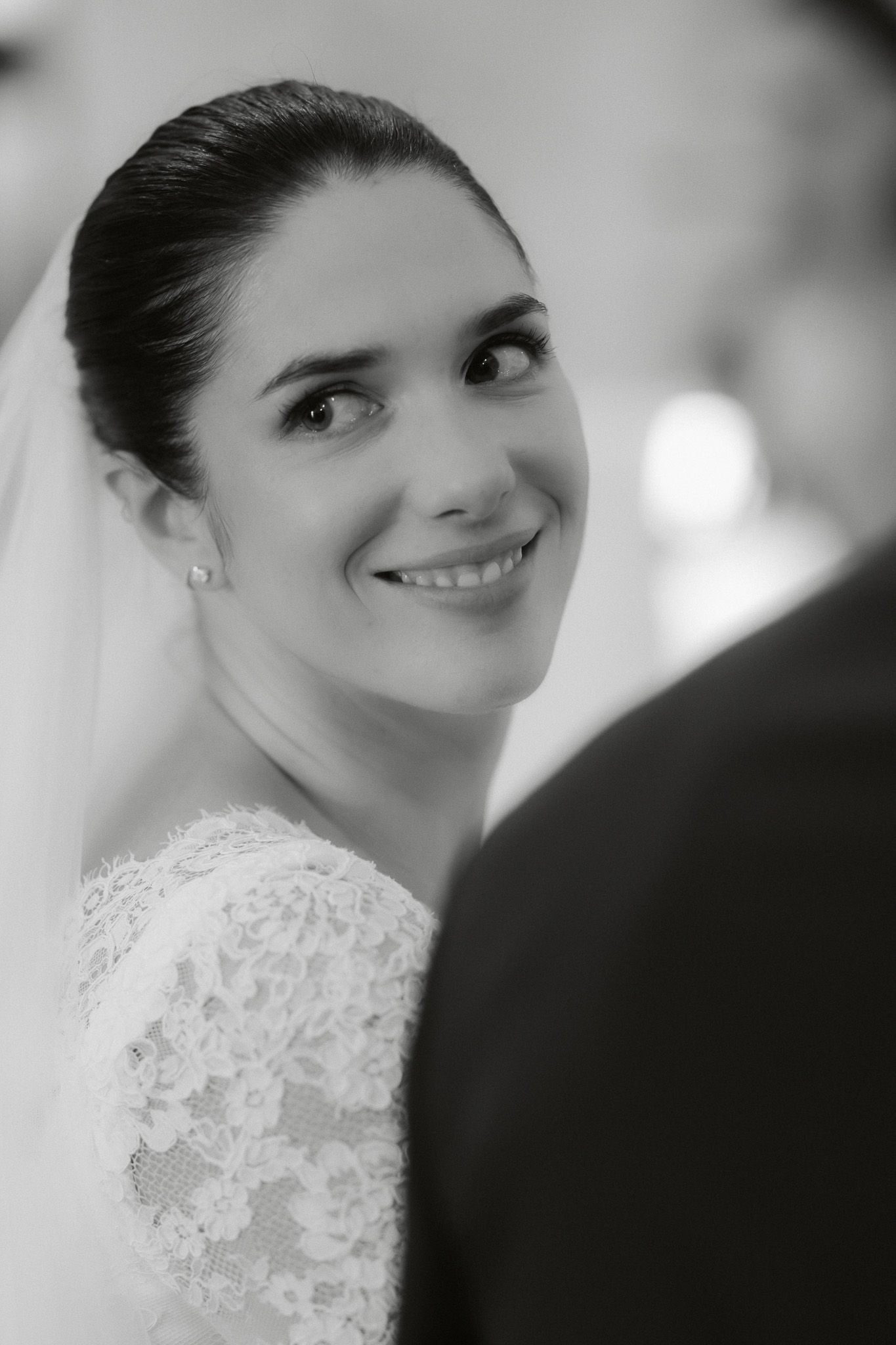 Black and white portrait of a smiling bride in lace dress and veil, gazing up at groom during wedding ceremony.