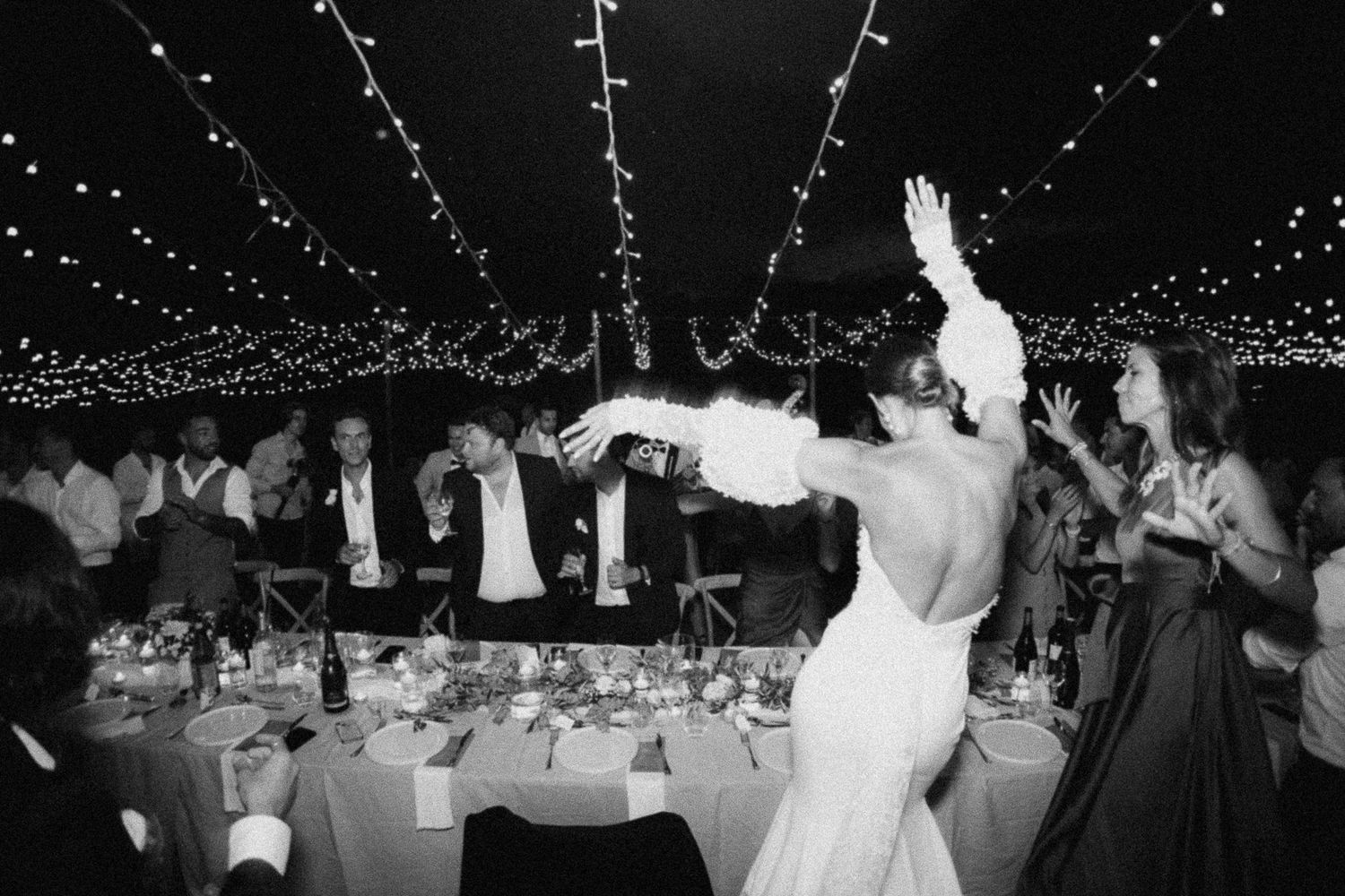 Black and white wedding reception photo of bride dancing under twinkling fairy lights with guests celebrating in background.