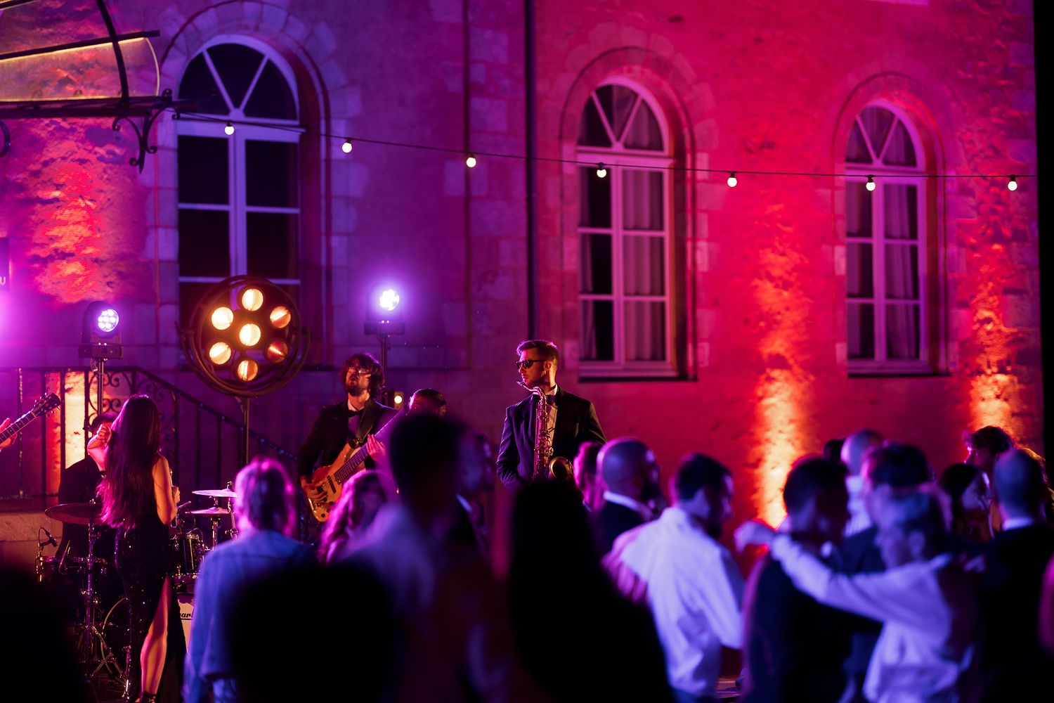 Live band performing outdoors at night with red and purple uplighting against a stone building with dancing guests.