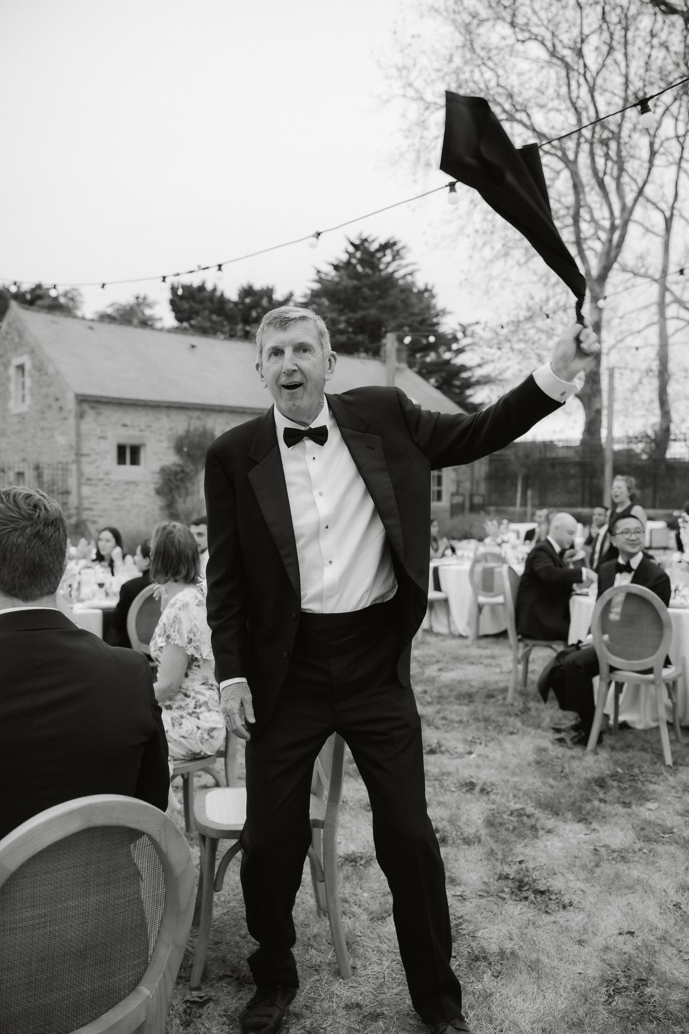 Man in tuxedo joyfully raises his hat at an outdoor wedding reception in black and white photo.