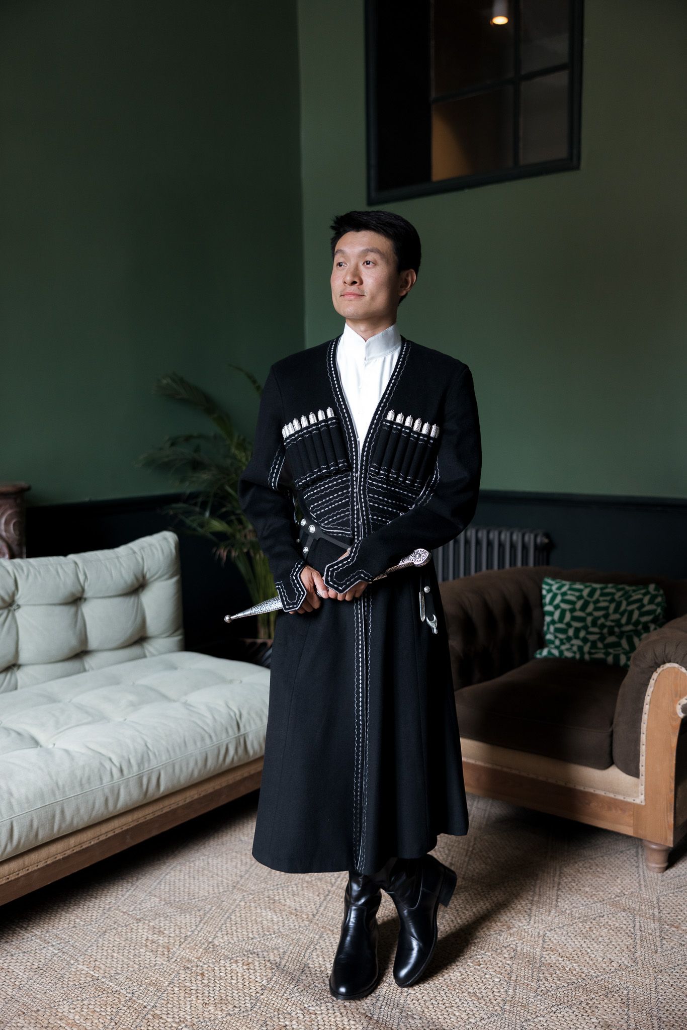 Man in traditional black Chokha coat with white embroidery holding a dagger, standing in elegant green interior.