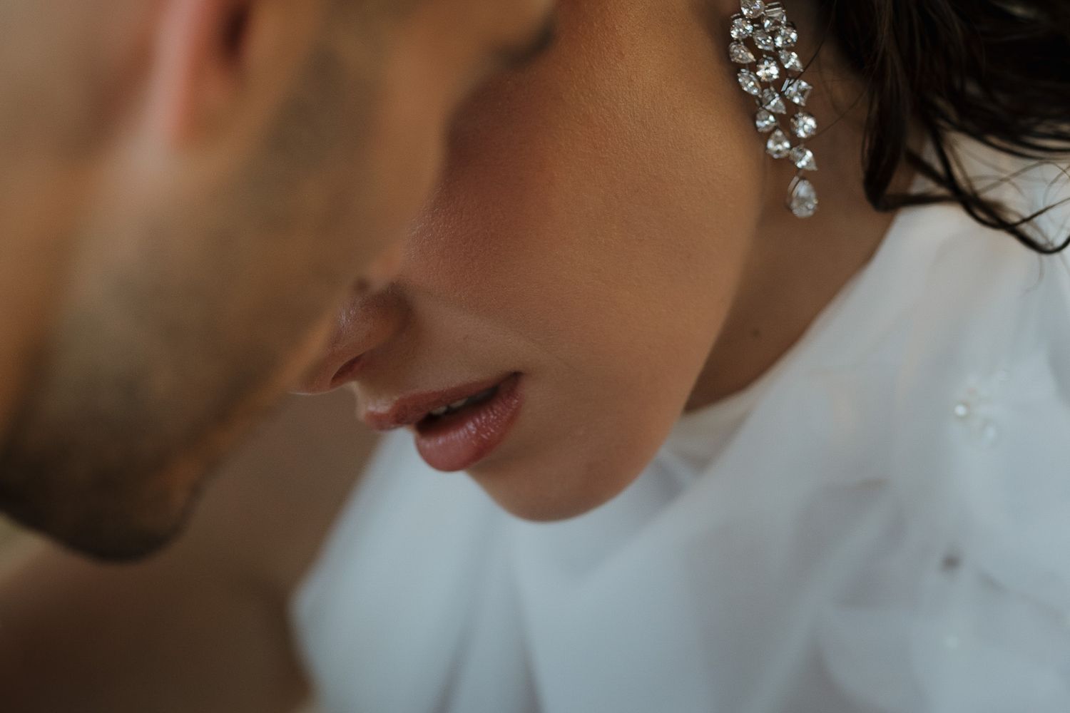 Close-up of a bride wearing sparkling diamond drop earrings in a white gown, with a groom leaning in close.