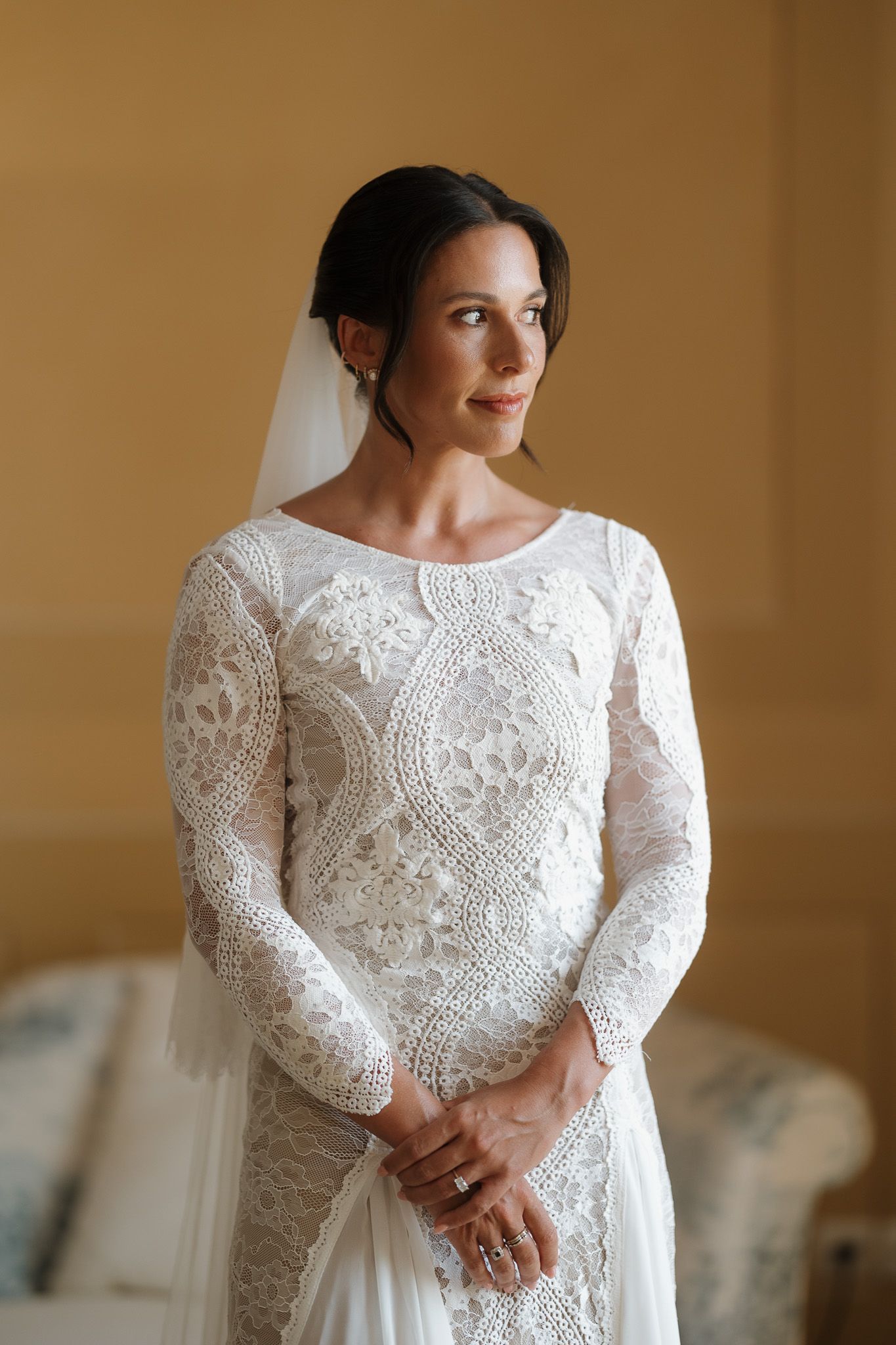 Elegant bride in long-sleeve white lace wedding dress with veil, posing indoors with a serene expression.
