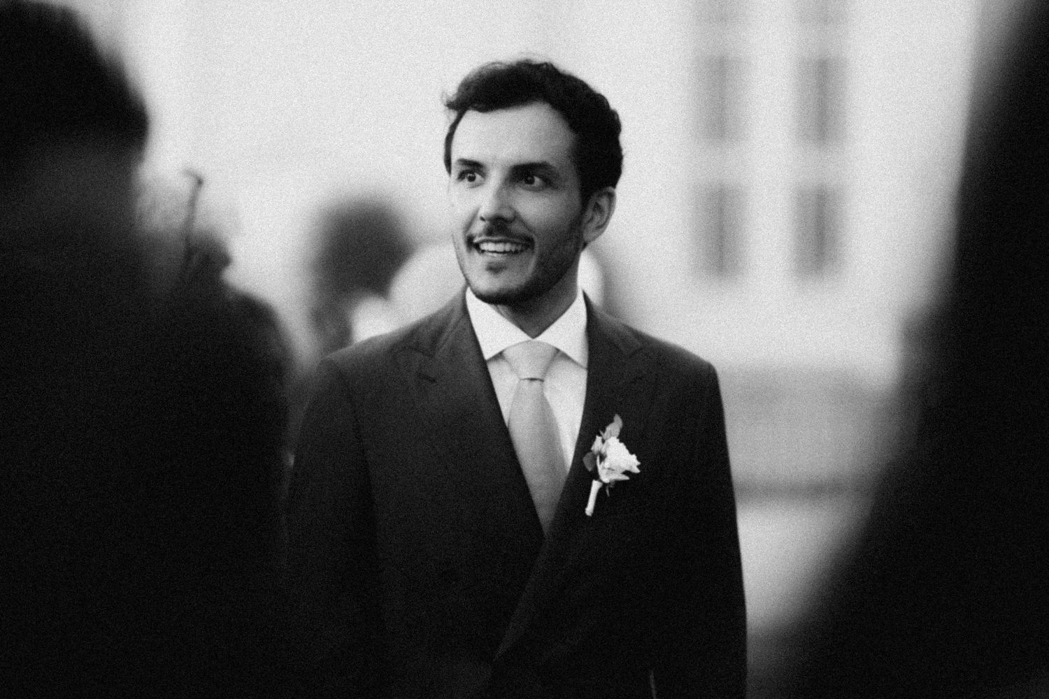 Black and white photo of a smiling groom in a suit with boutonniere at a wedding ceremony.