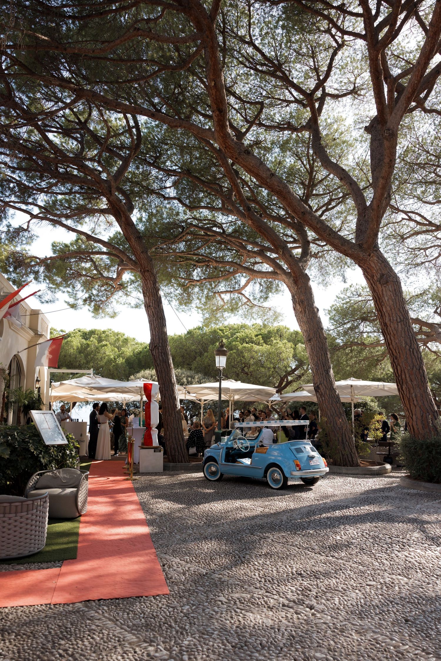 A vintage blue Fiat 500 parked under tall pine trees at an elegant outdoor event venue with red carpet and white umbrellas.