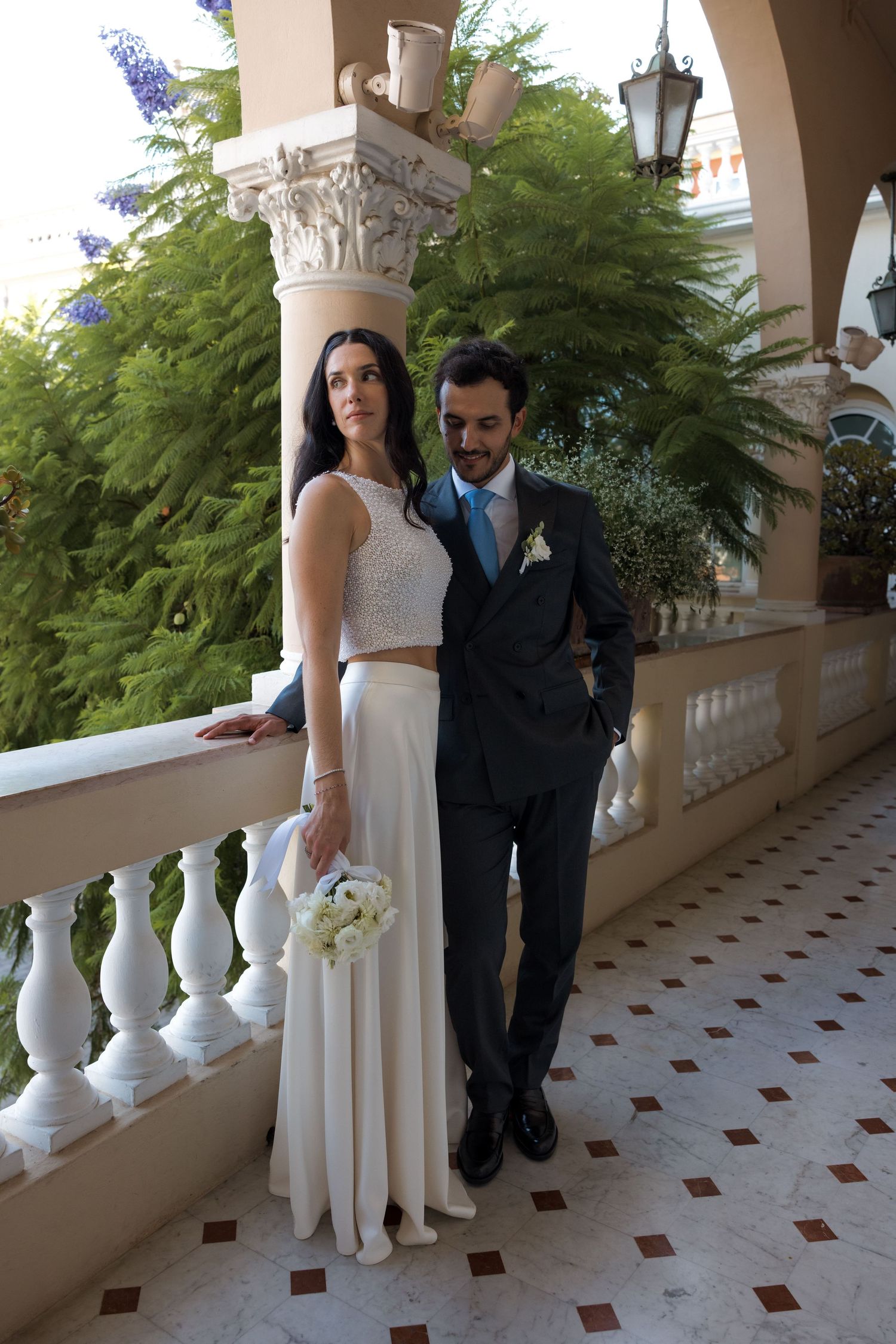 Bride and groom posing on an elegant balcony with white railings and columns, surrounded by lush greenery.