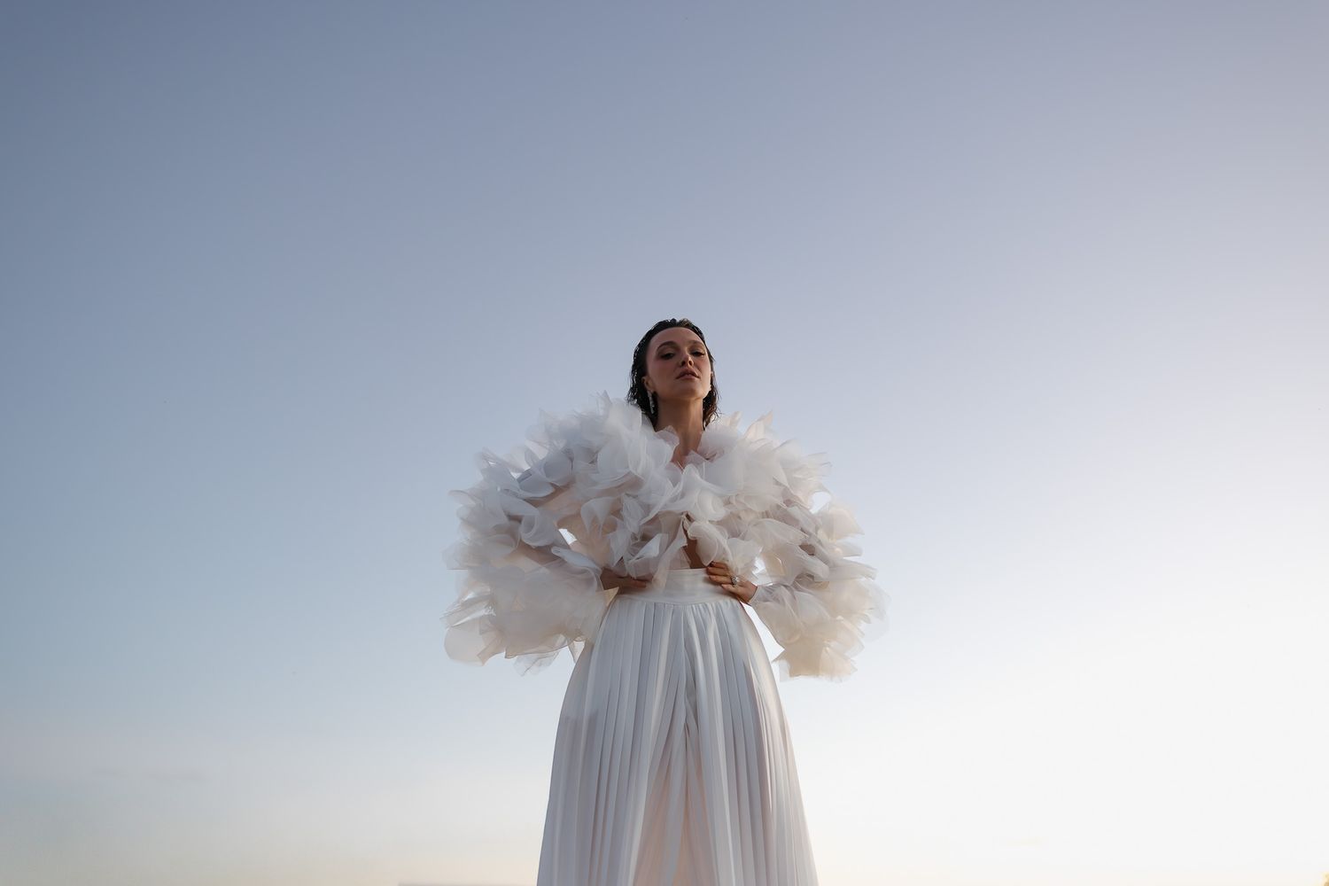 Woman in white pleated dress with feathered cape poses against soft blue sky backdrop.