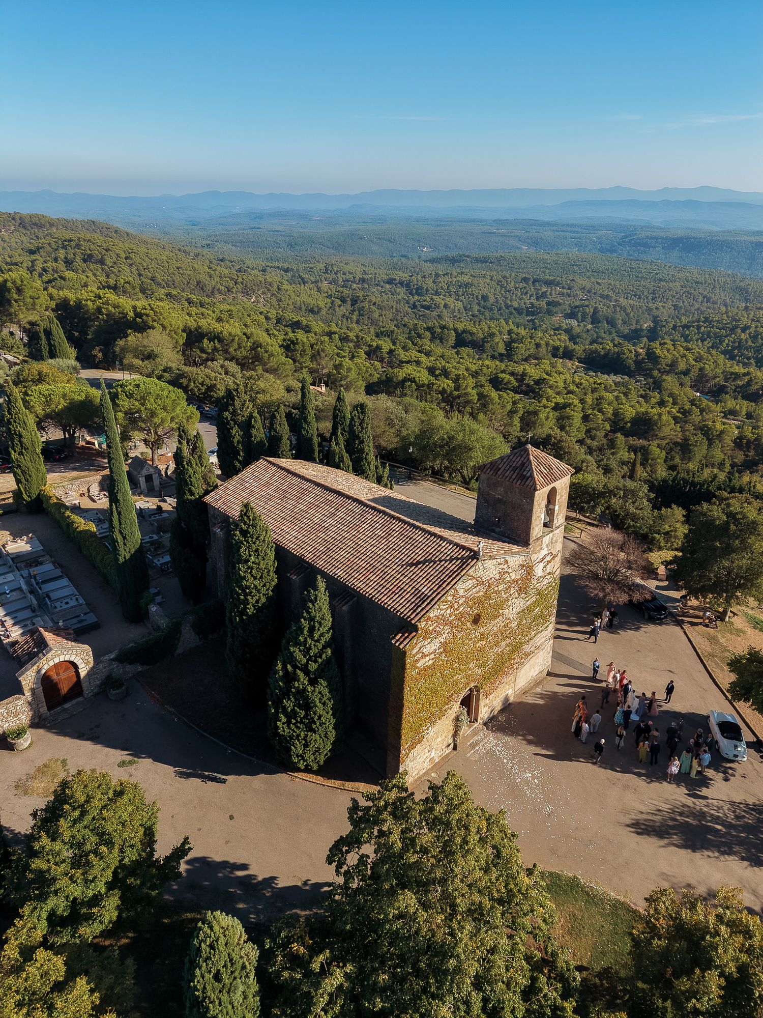 Aerial view of a historic stone chapel surrounded by cypress trees atop a hill with vast green forest landscape.