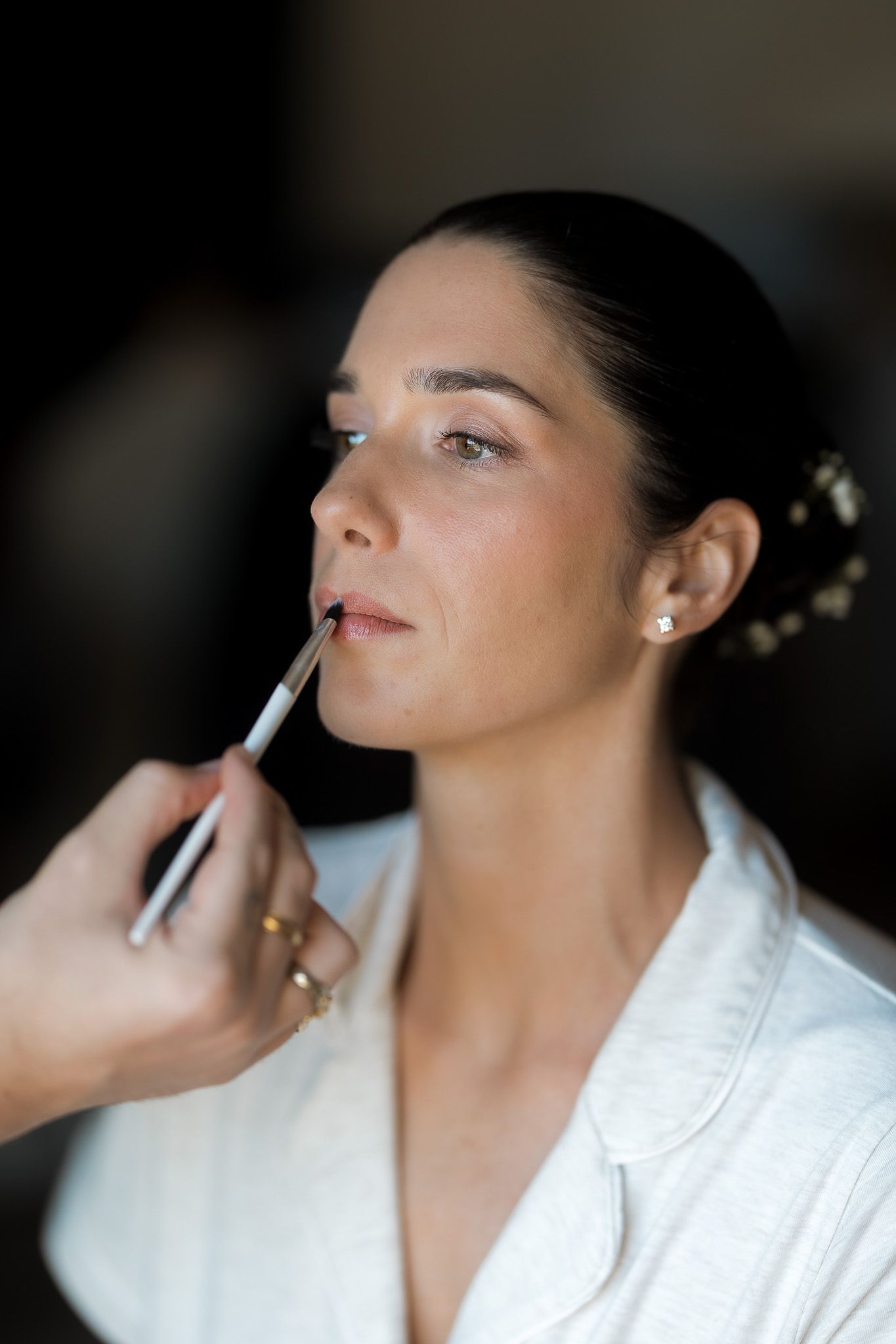 A makeup artist applies lip liner to a brunette woman in a white robe, preparing her for a special occasion.
