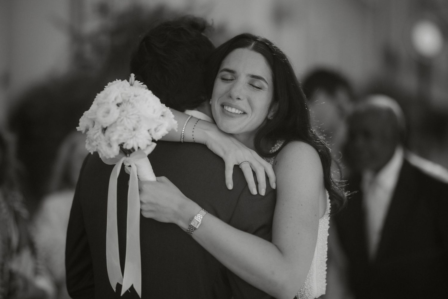 Black and white wedding photo of smiling bride hugging groom while holding white flower bouquet with ribbon.