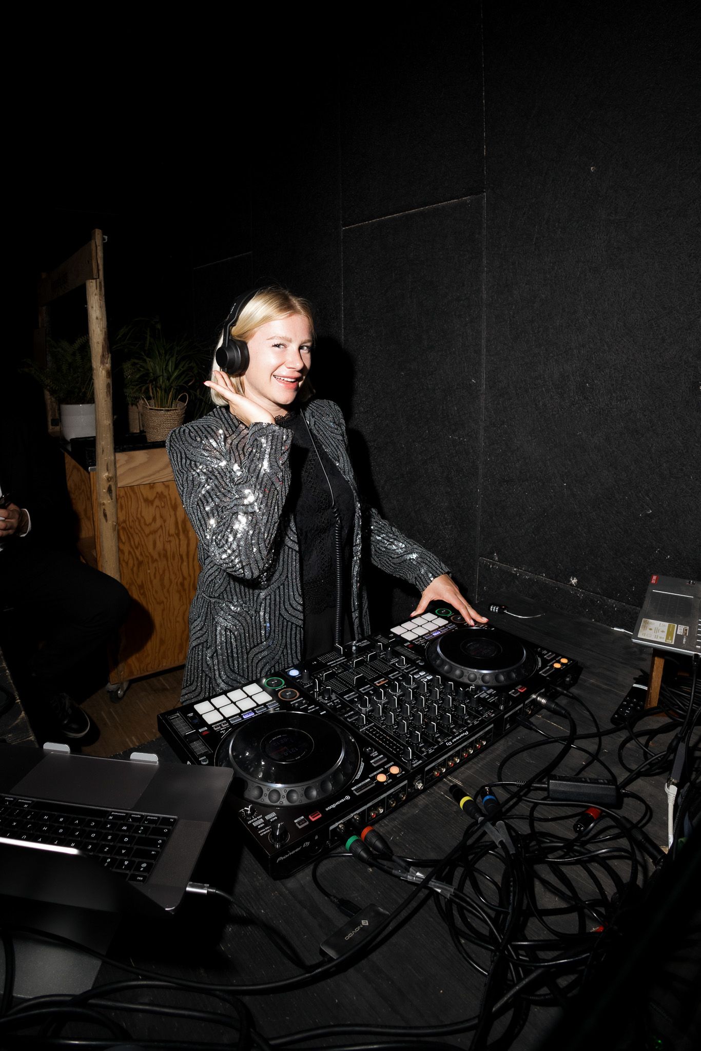 Smiling female DJ in sequin jacket mixing music on Pioneer DJ controller while wearing headphones at a dark venue.