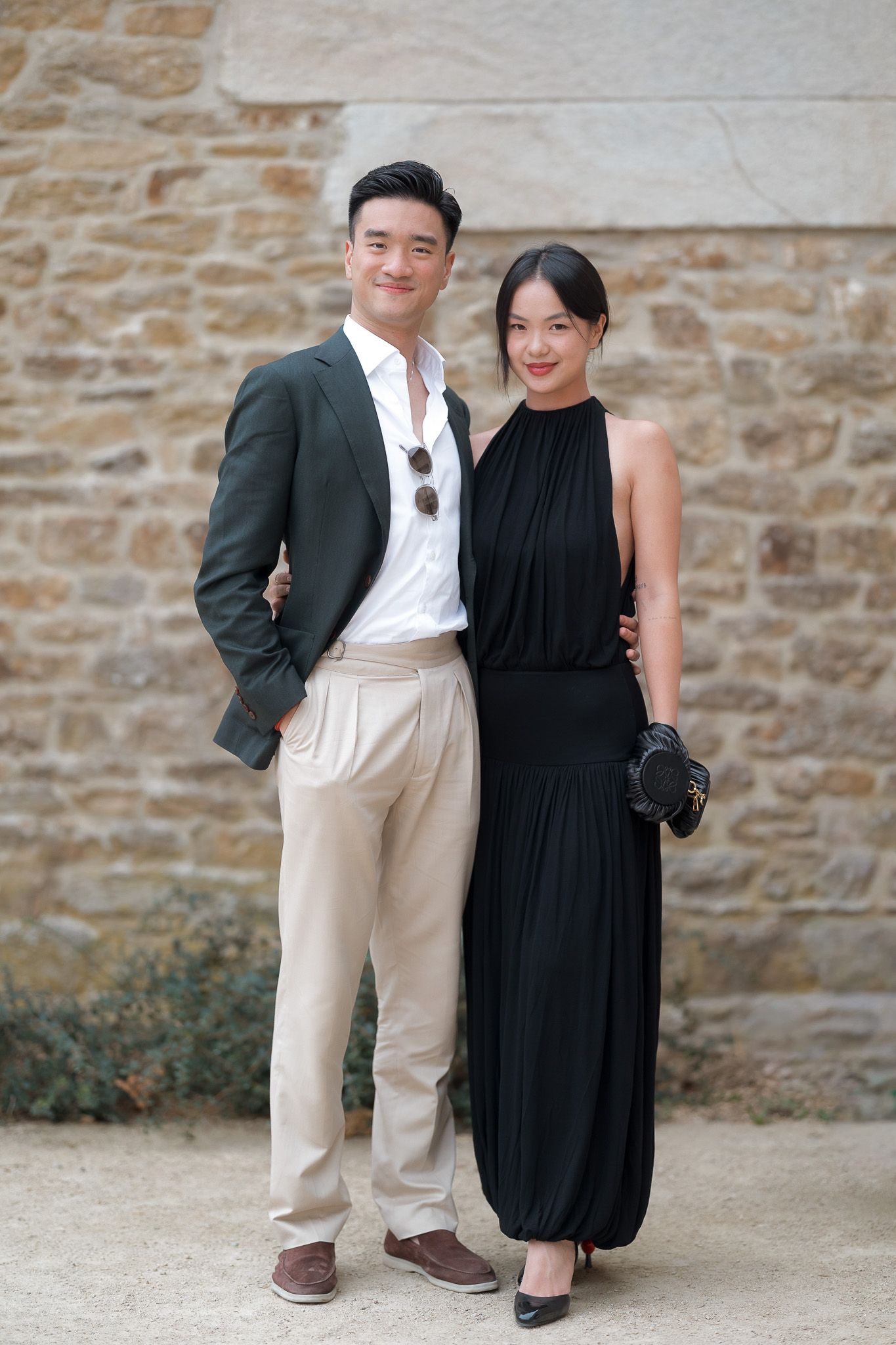 Elegant couple posing together, man in dark blazer and woman in black halter maxi dress, standing before a stone wall.