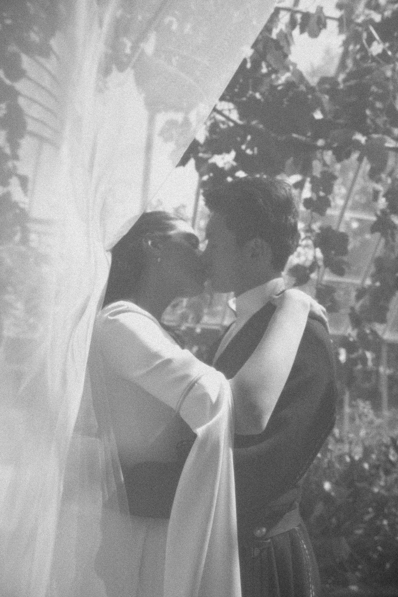 Black and white photo of a couple sharing a romantic kiss under a sheer veil outdoors near lush green foliage.